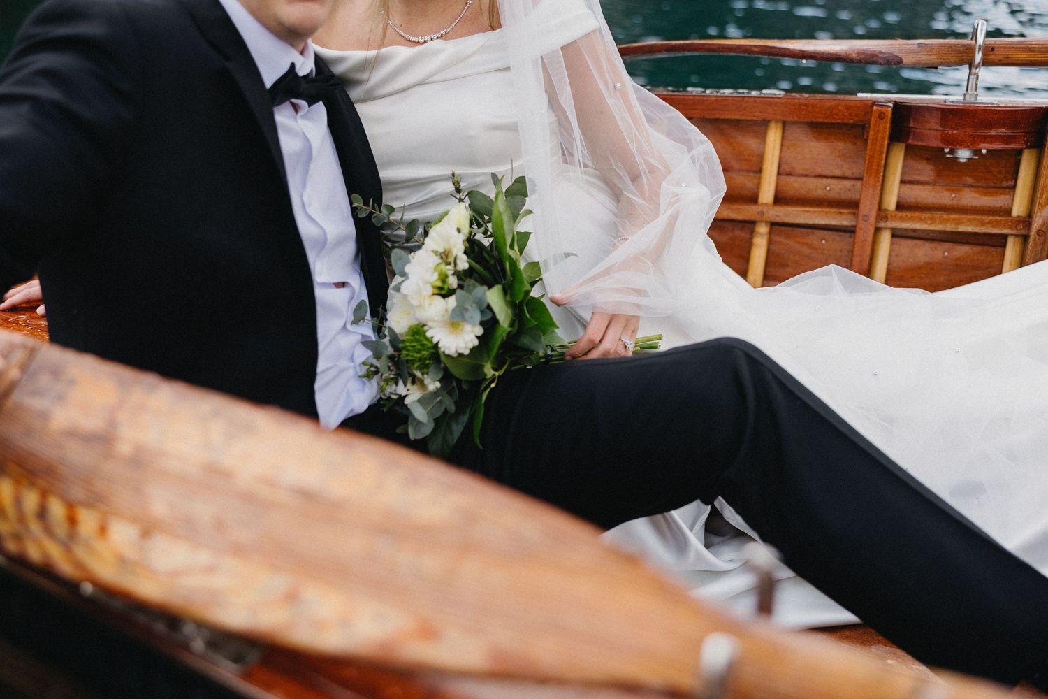 Close up of bride and groom sitting together in wooden boat with wedding attire details.