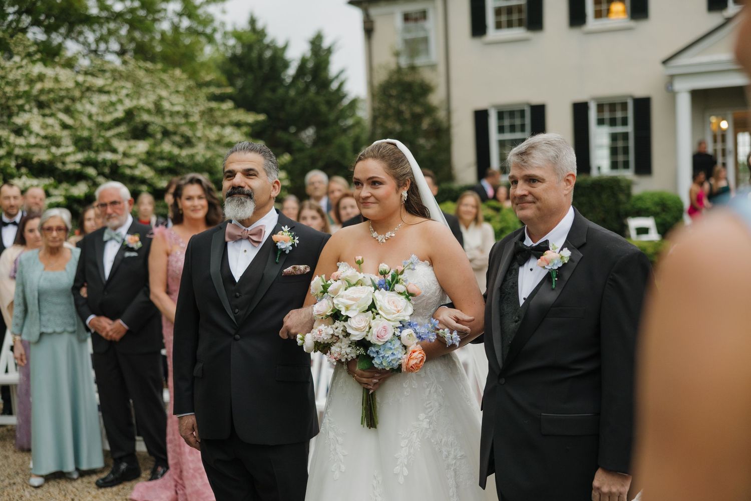 Bride in white gown walks down outdoor aisle escorted by two men in tuxedos, holding pastel bouquet at elegant estate wedding.