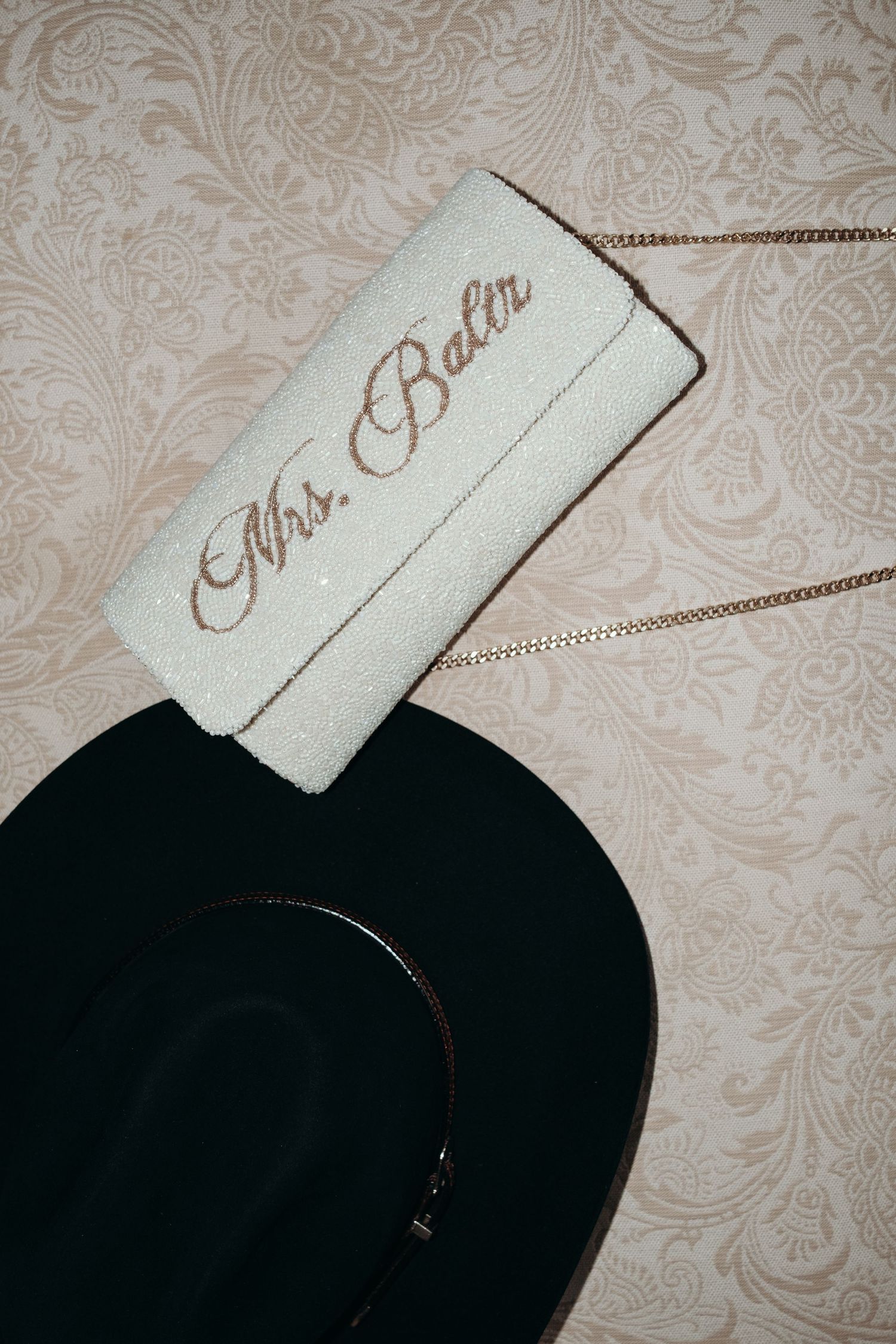 White embroidered Mrs. Baltr clutch purse with gold chain strap resting on black wide-brim hat on floral wallpaper.