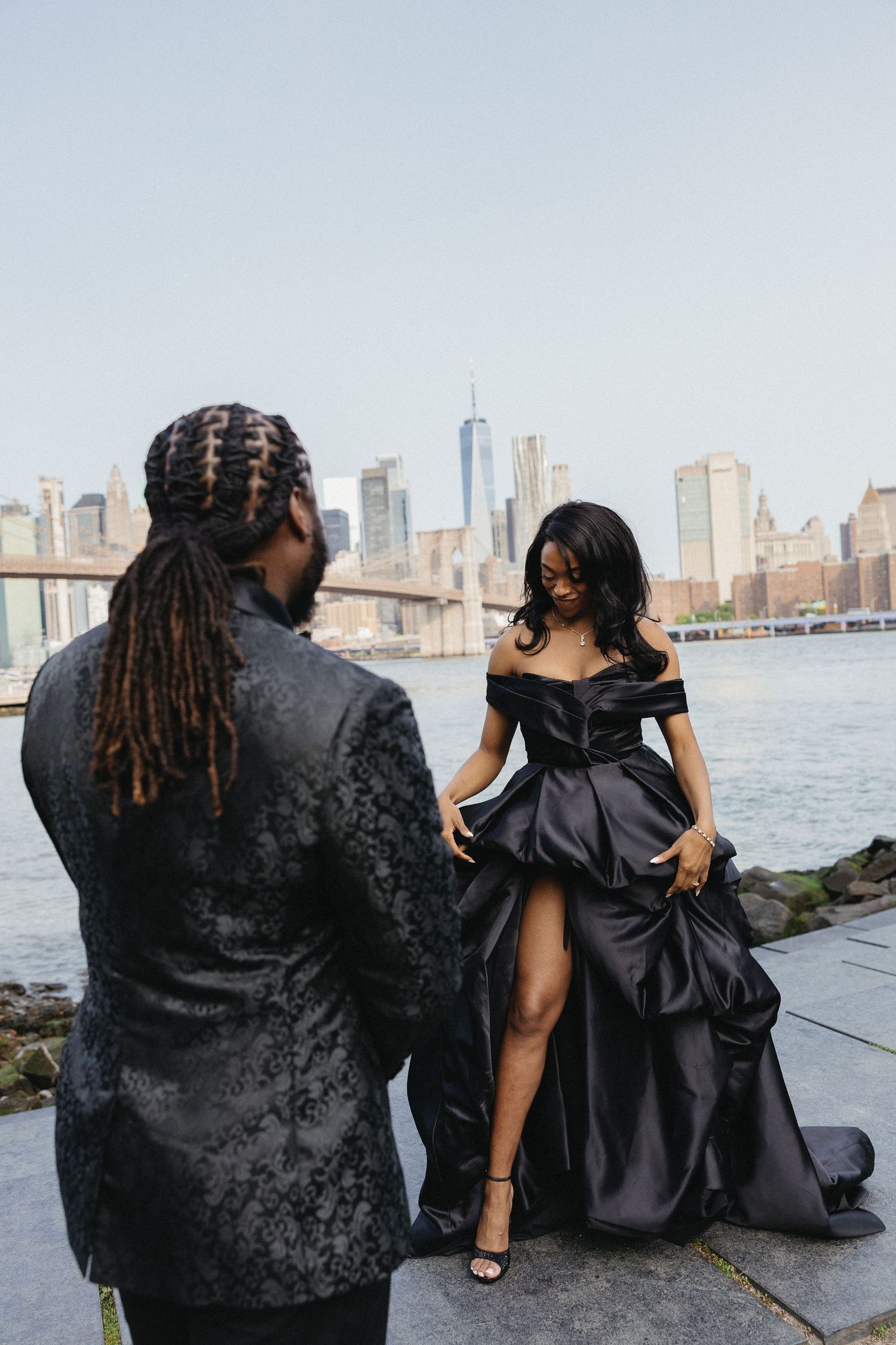 Couple in formal wear shares romantic moments by the water with Manhattan skyline behind them.