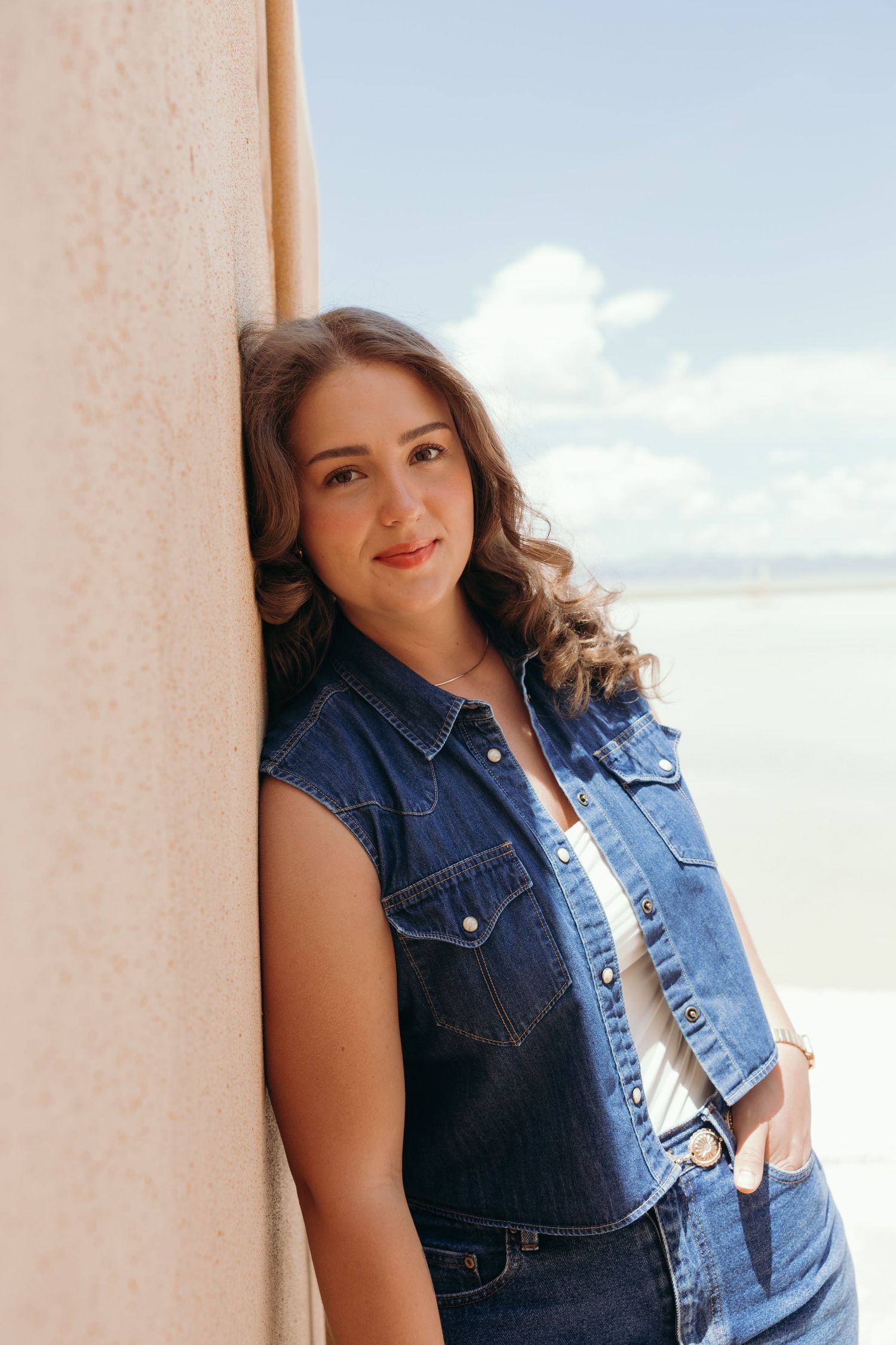 A woman in a denim vest poses against a light-colored wall with a bright sky in the background.
