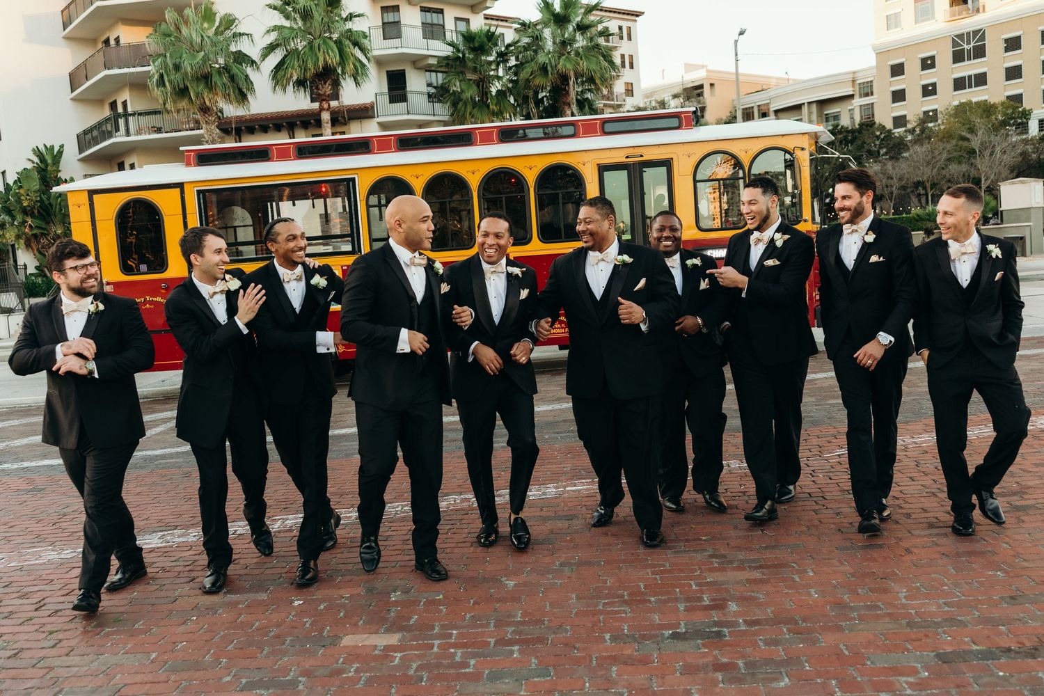 A long line of wedding party members in black suits dance in a choreographed sequence outdoors at a reception.
