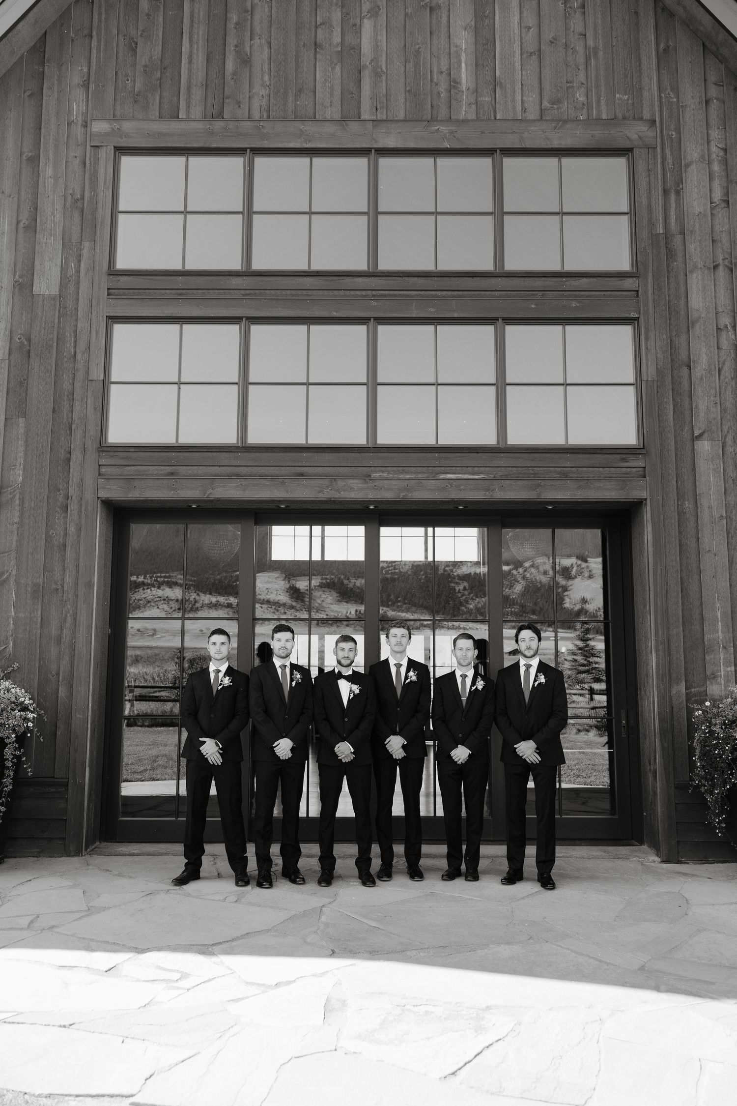 Wedding party in black suits poses in front of a rustic wooden barn with large windows.