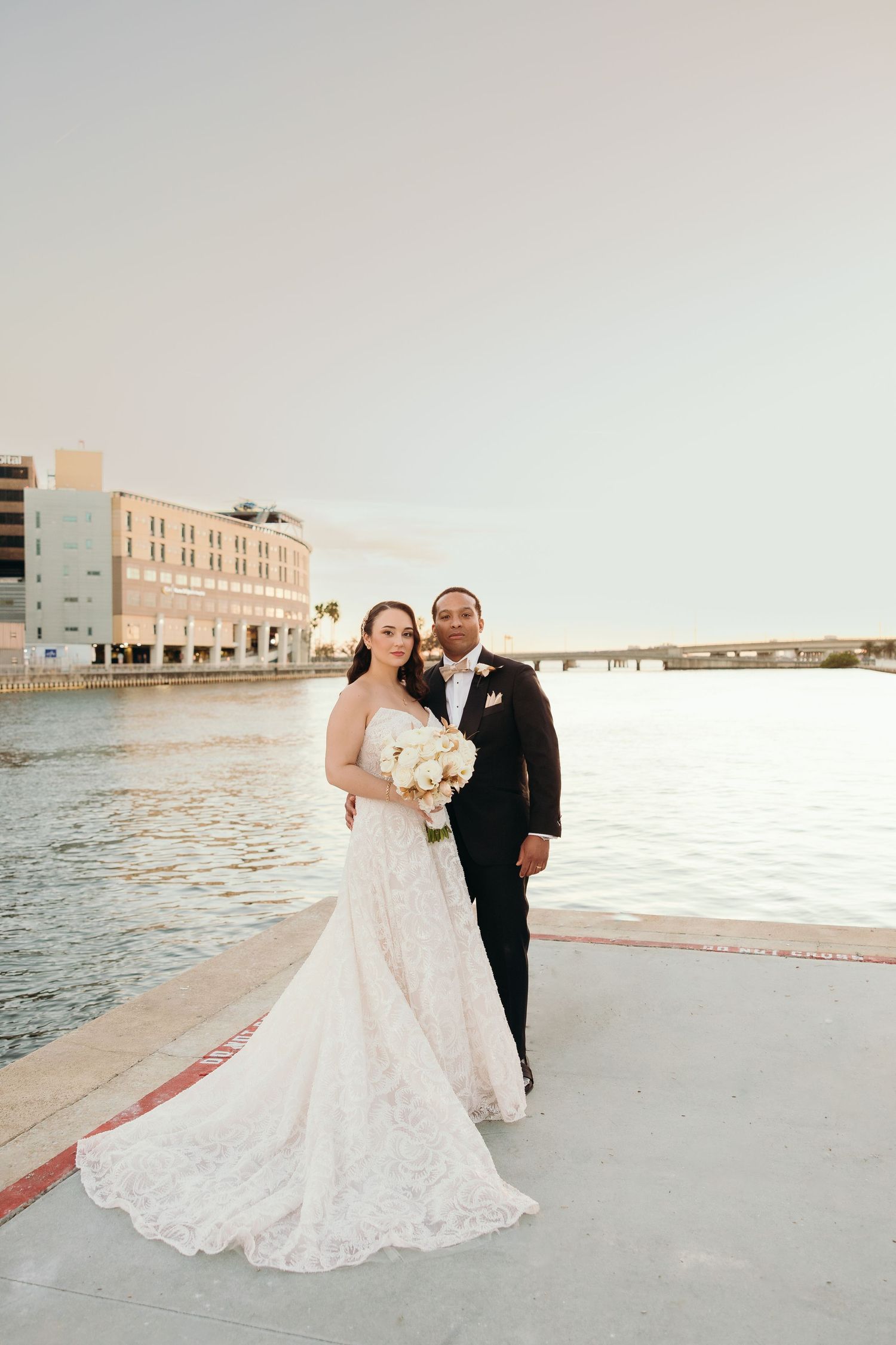 A newlywed couple poses together on a waterfront with a beautiful sunset and urban backdrop.