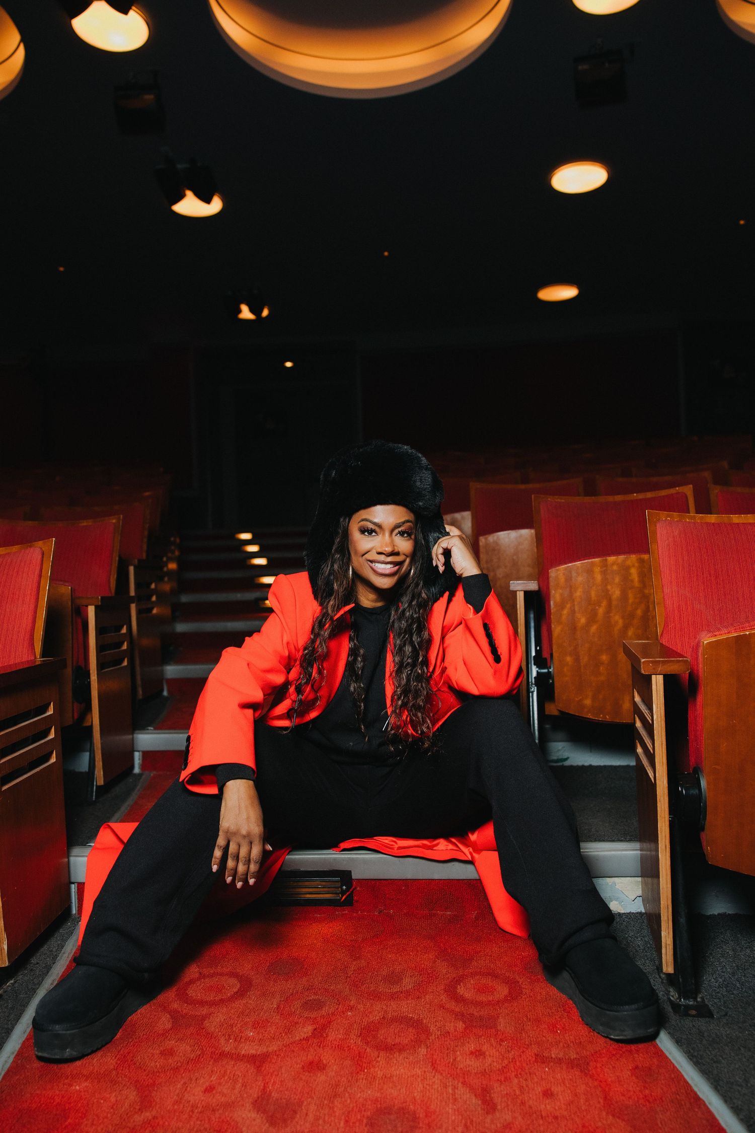 Woman in red coat and black beret sits confidently in theater seats, smiling at camera.