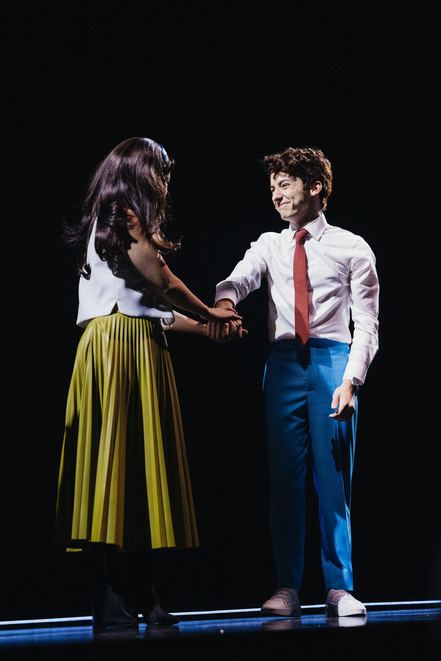 Two performers on a dark stage, one wearing a yellow skirt and another in blue pants with a red tie.