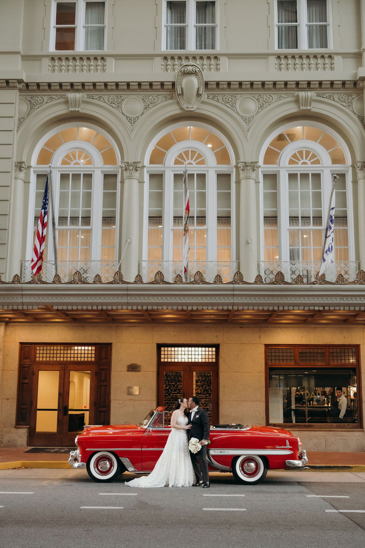A classic red convertible car parked in front of a historic building with arched windows and American flag.