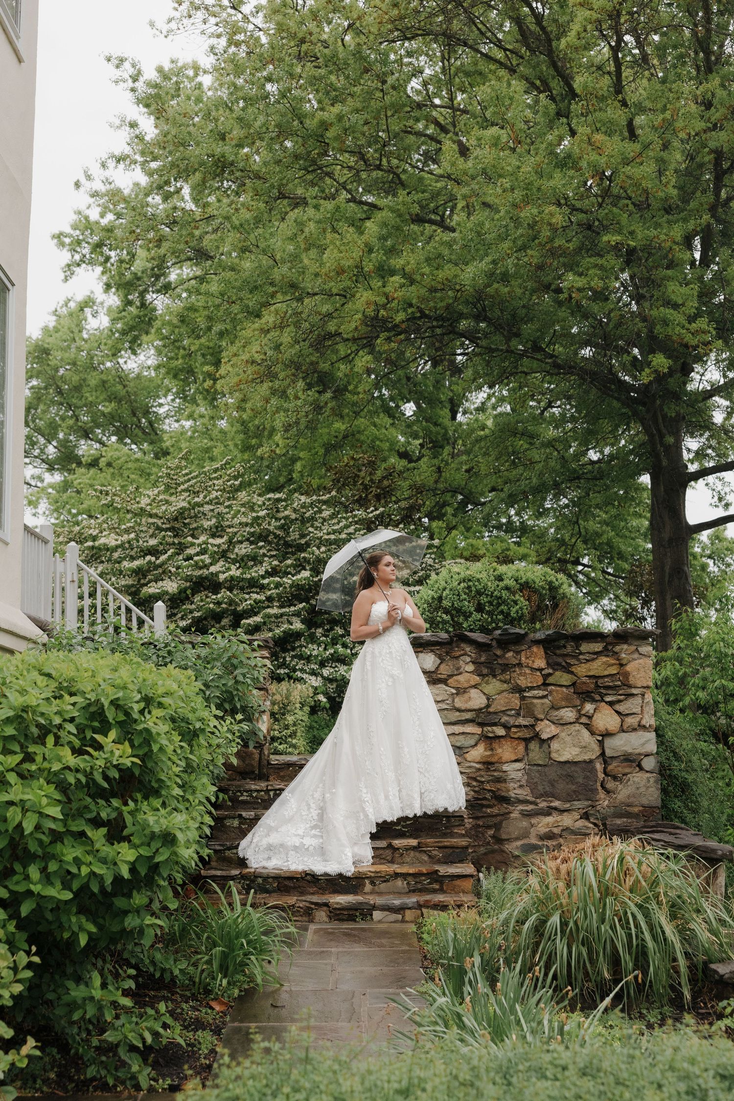 Bride in white lace gown holding parasol umbrella stands on stone steps surrounded by lush green garden foliage.