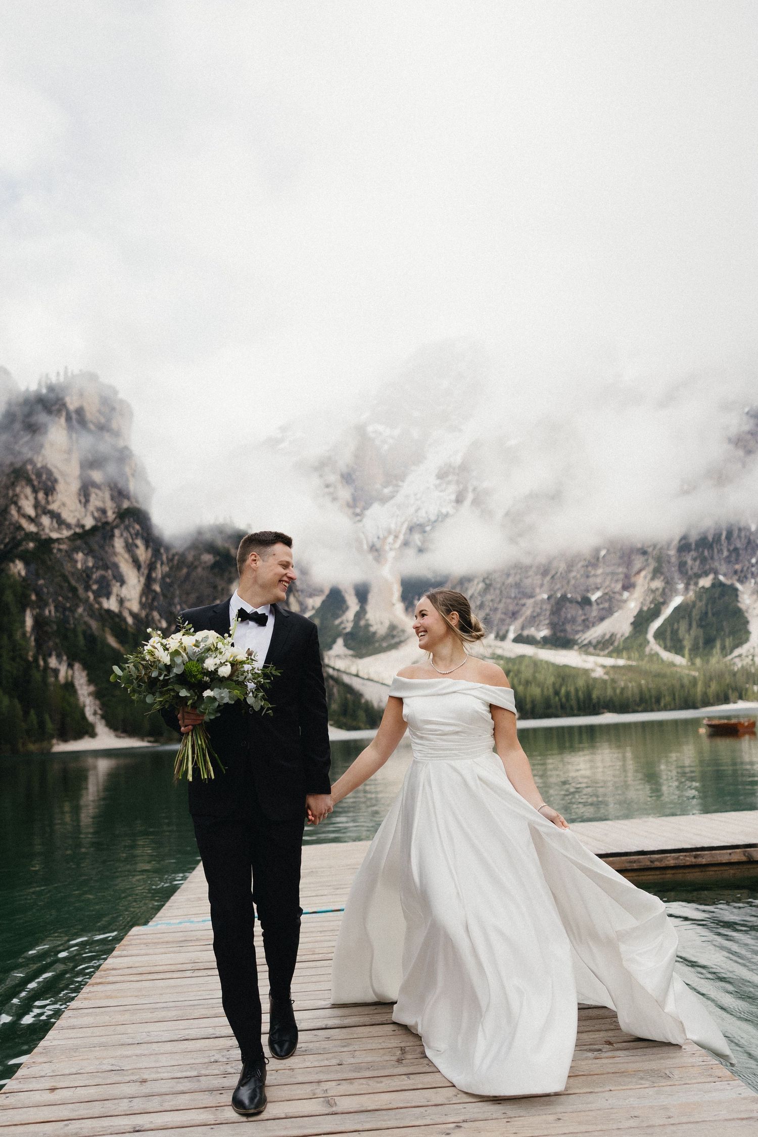 Newlyweds walking on a dock by a mountain lake with the bride's dress flowing in the wind.