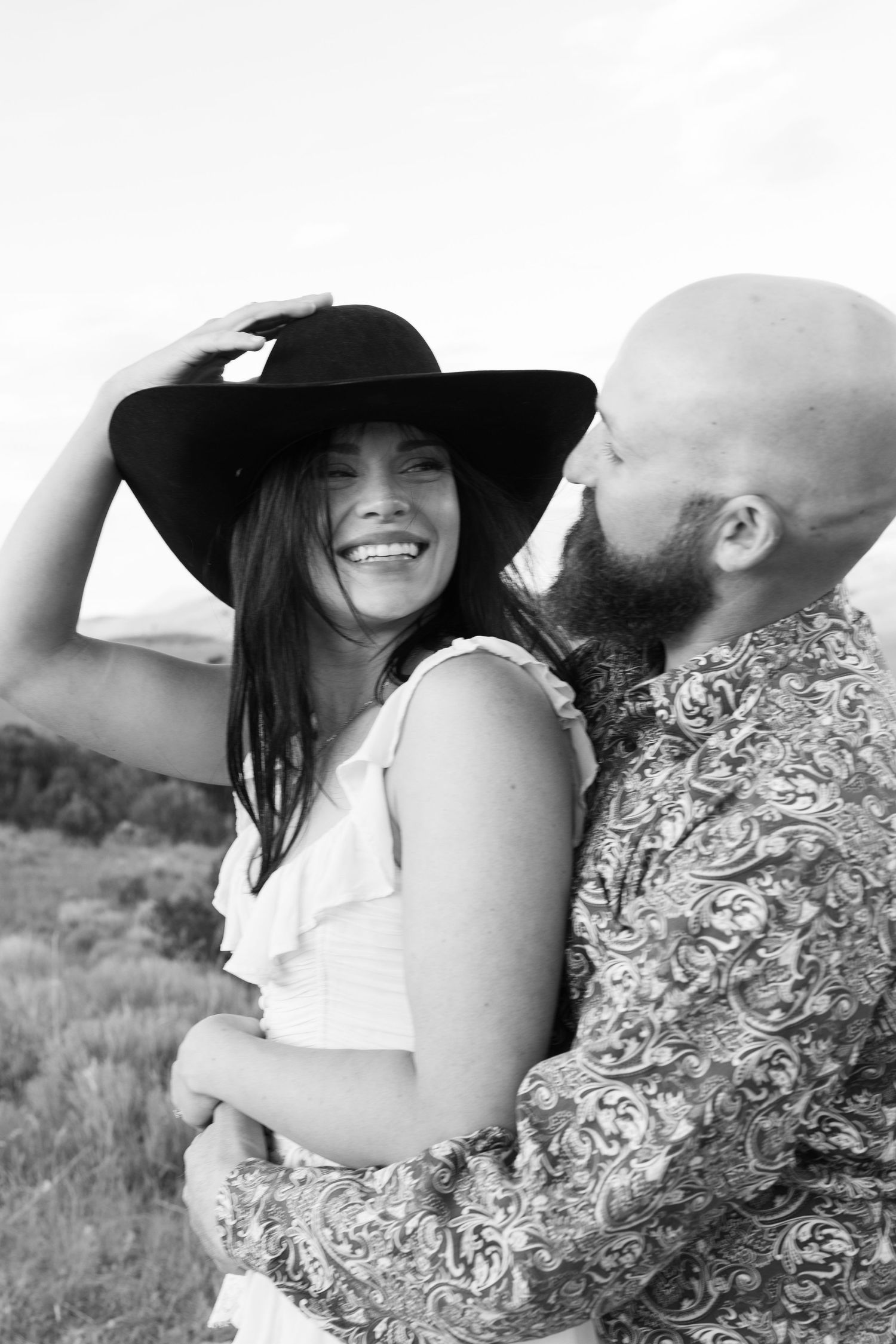 Couple in western attire embracing in a field with mountains in the background.