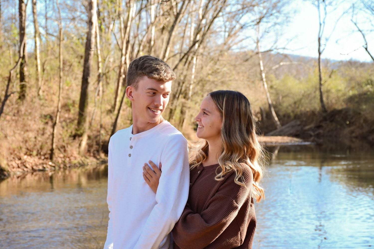 A young couple posing by a river with the man facing opposite of her but also admiring her from the front, while she holds onto him.