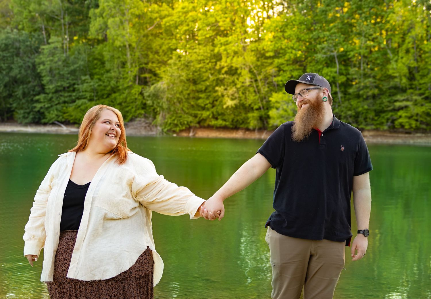 A newly engaged couple posing by reaching their hands out to each other in front of a lake.