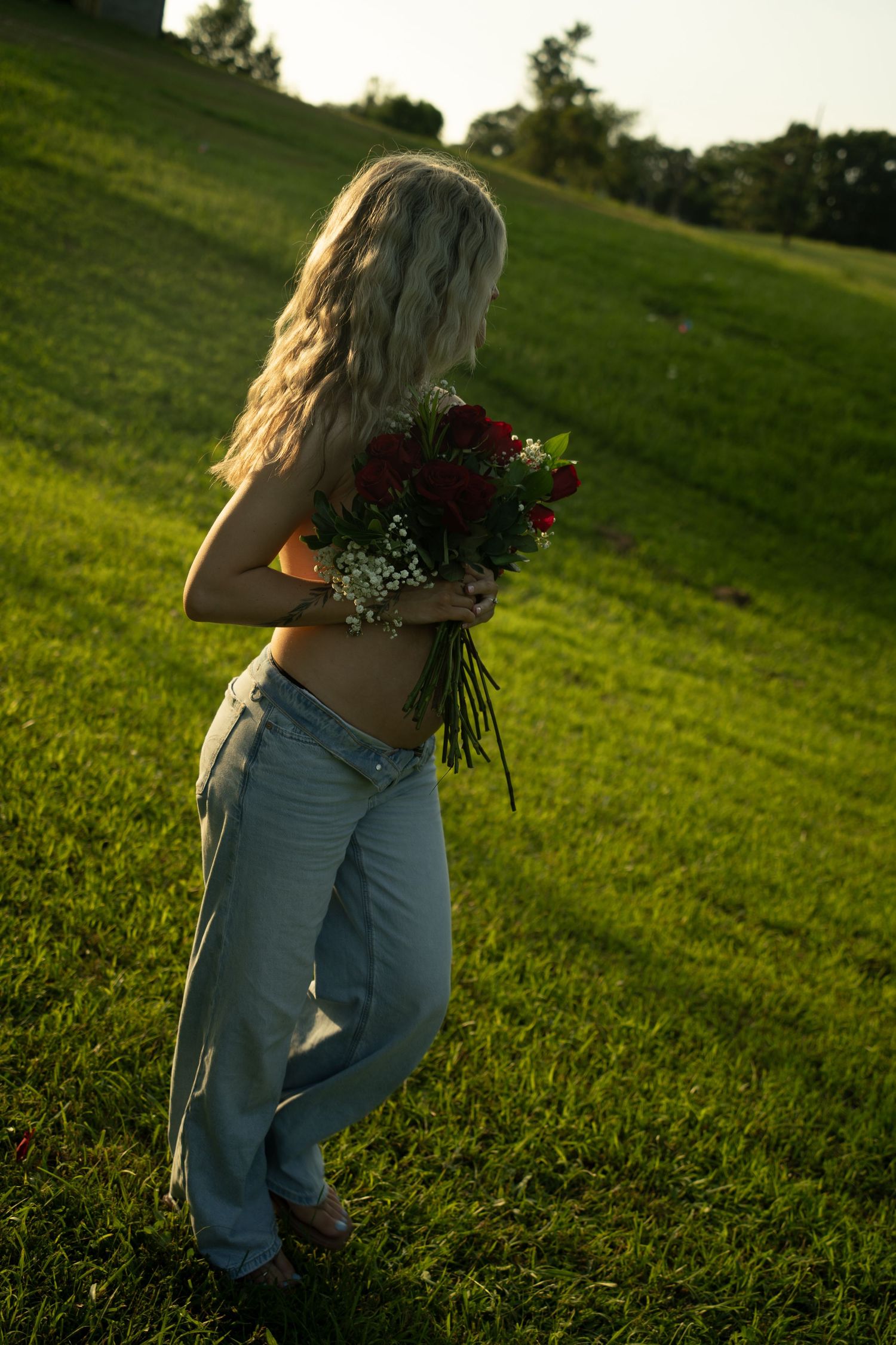 Young woman with long blonde hair holding red roses bouquet in green park at golden hour sunset.