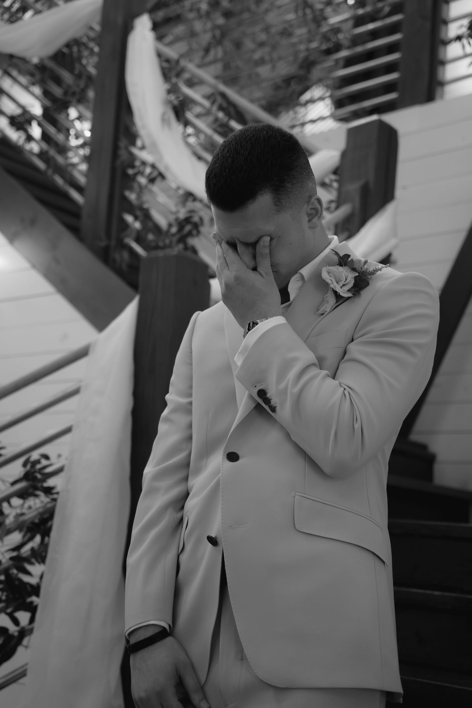Emotional groom in white suit wiping tears during wedding ceremony outdoors.