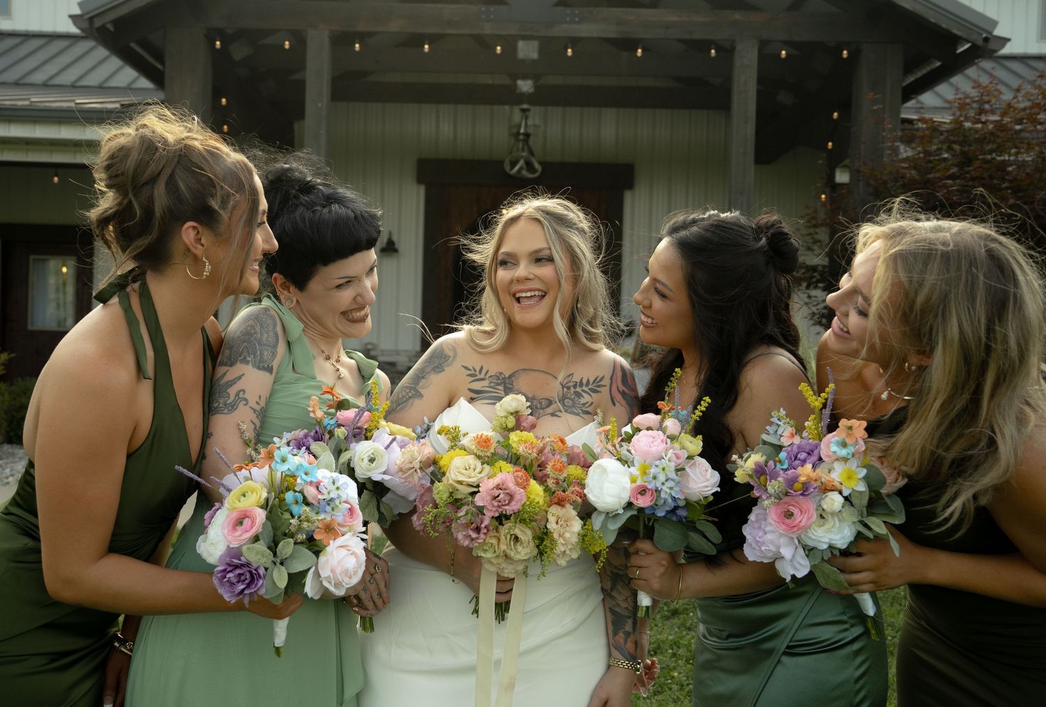 Bride in white gown laughing with four bridesmaids in green dresses holding colorful bouquets outdoors.