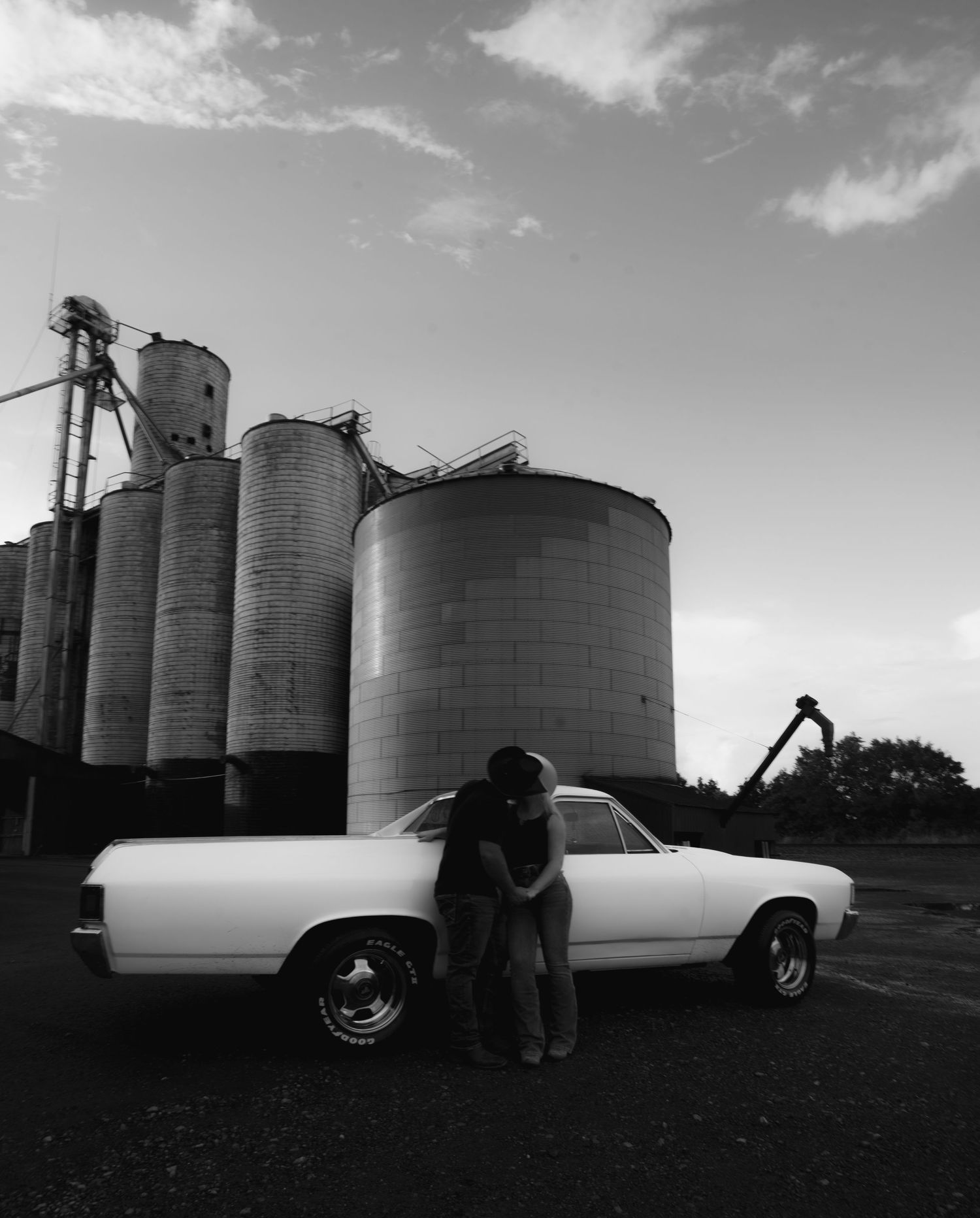 Couple embracing beside a classic white car in front of large grain silos, black and white rural photography.