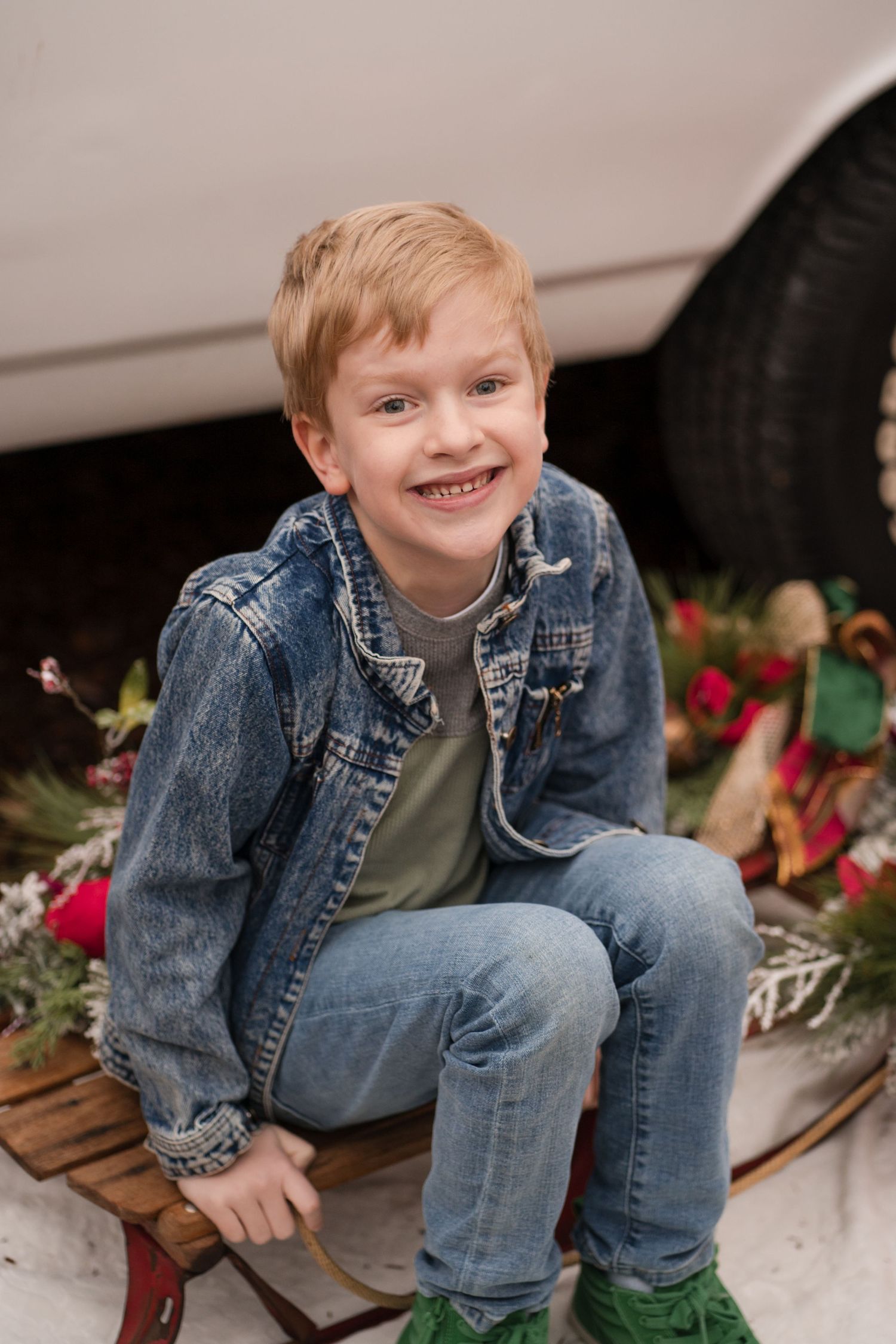 A child wearing a denim jacket and jeans sits on a wooden sled with holiday decorations and snow in the background.