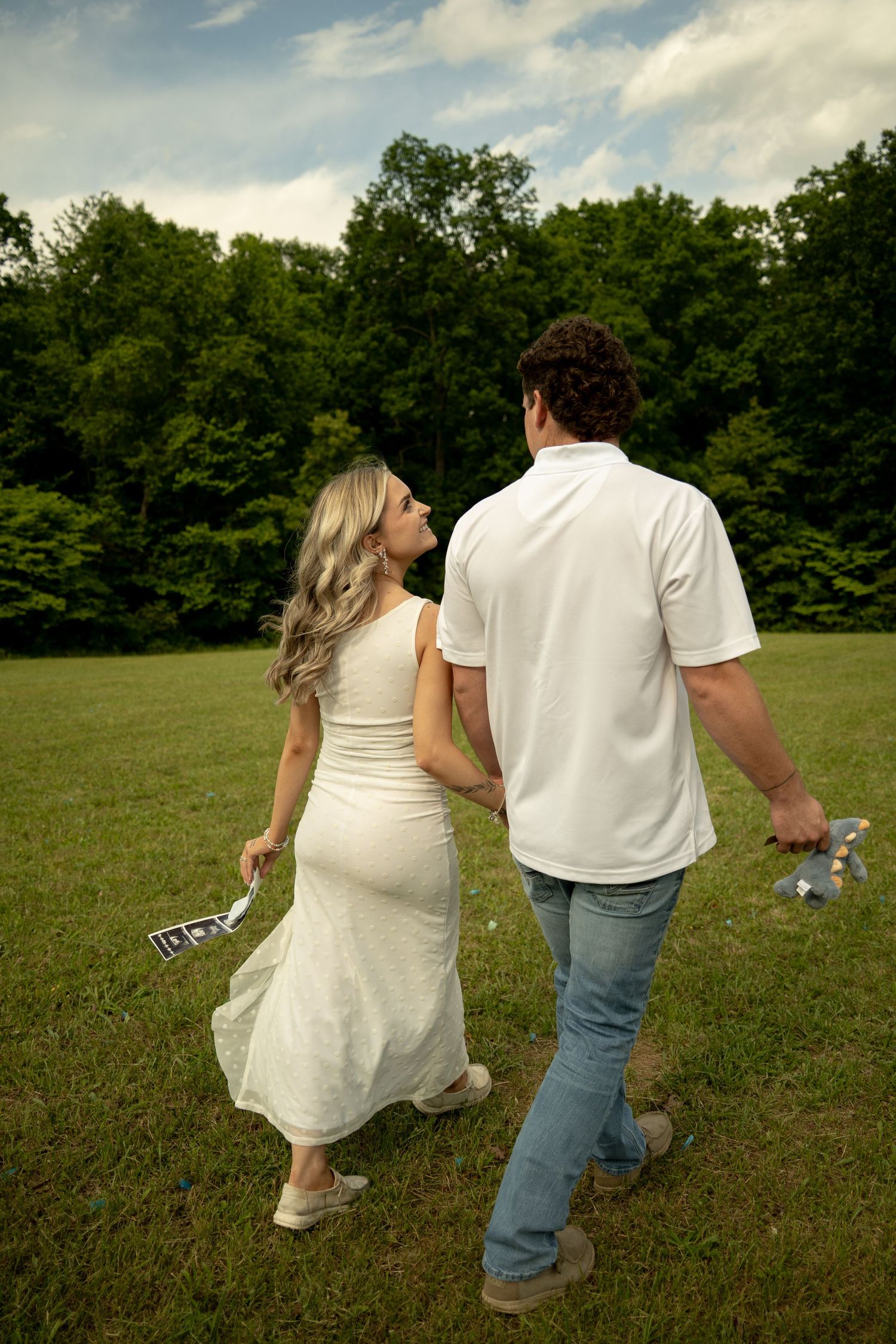 Pregnant couple walking hand-in-hand across green field holding ultrasound photos, woman in white dress, man in white shirt and jeans.
