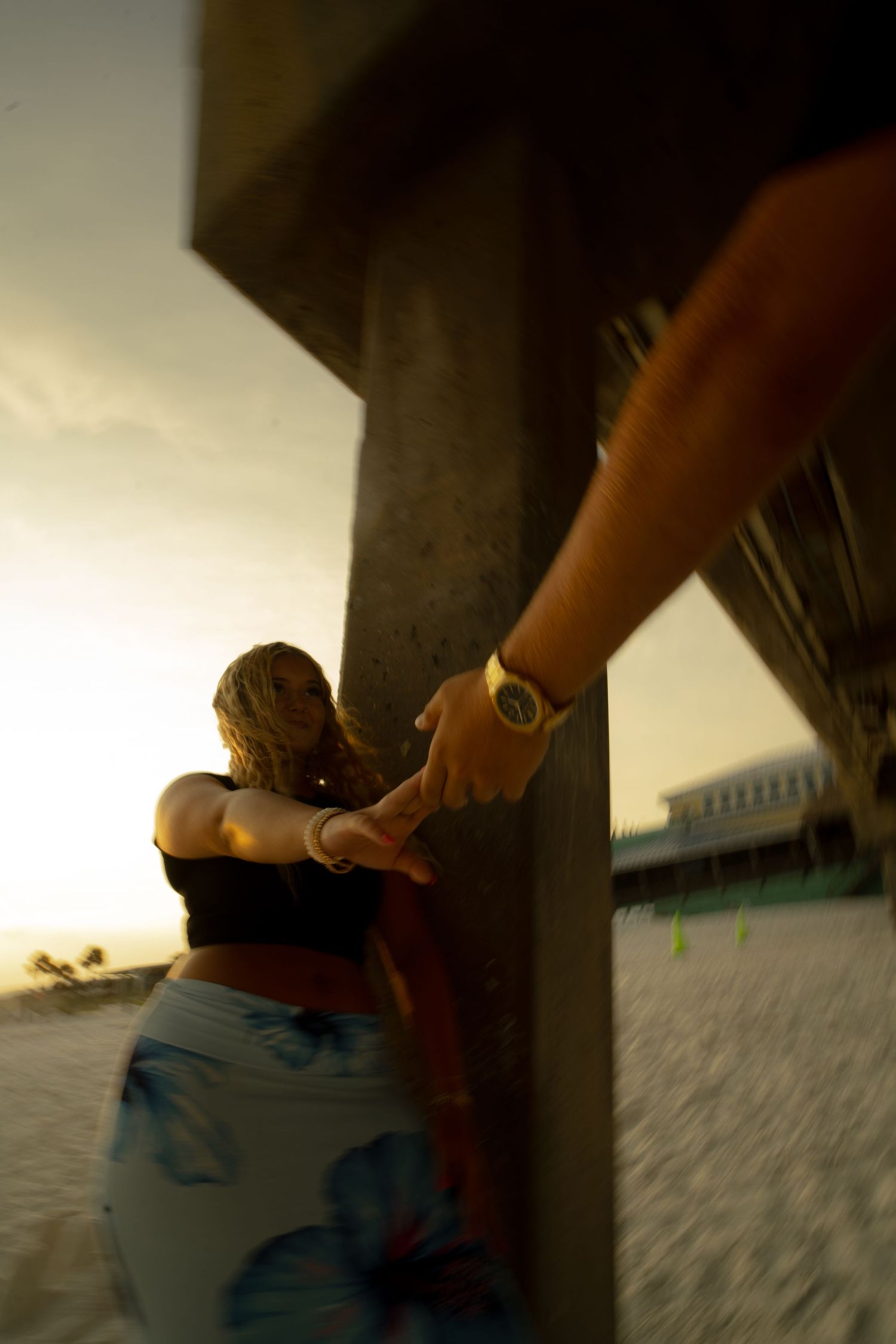 Couple holding hands on a sandy beach at sunset near a wooden pier, woman in floral skirt with golden hour glow.