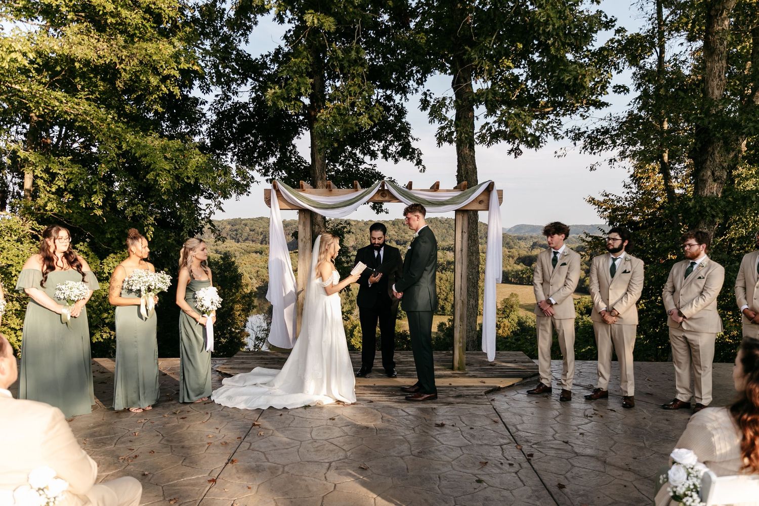 Wedding ceremony takes place on stone patio under wooden arch with wedding party in gray and beige attire.