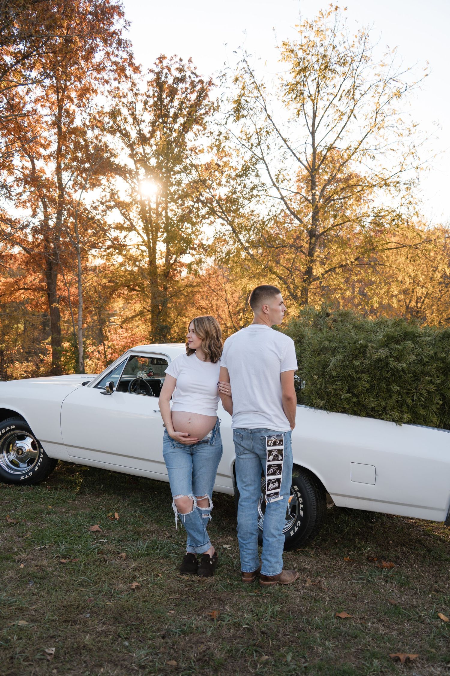 A couple in white and denim poses near a classic white car during a sunset photoshoot in an autumn setting.