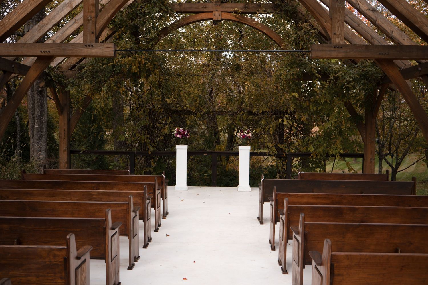 Empty wooden church pews under a rustic wooden arch in an outdoor wedding setting.