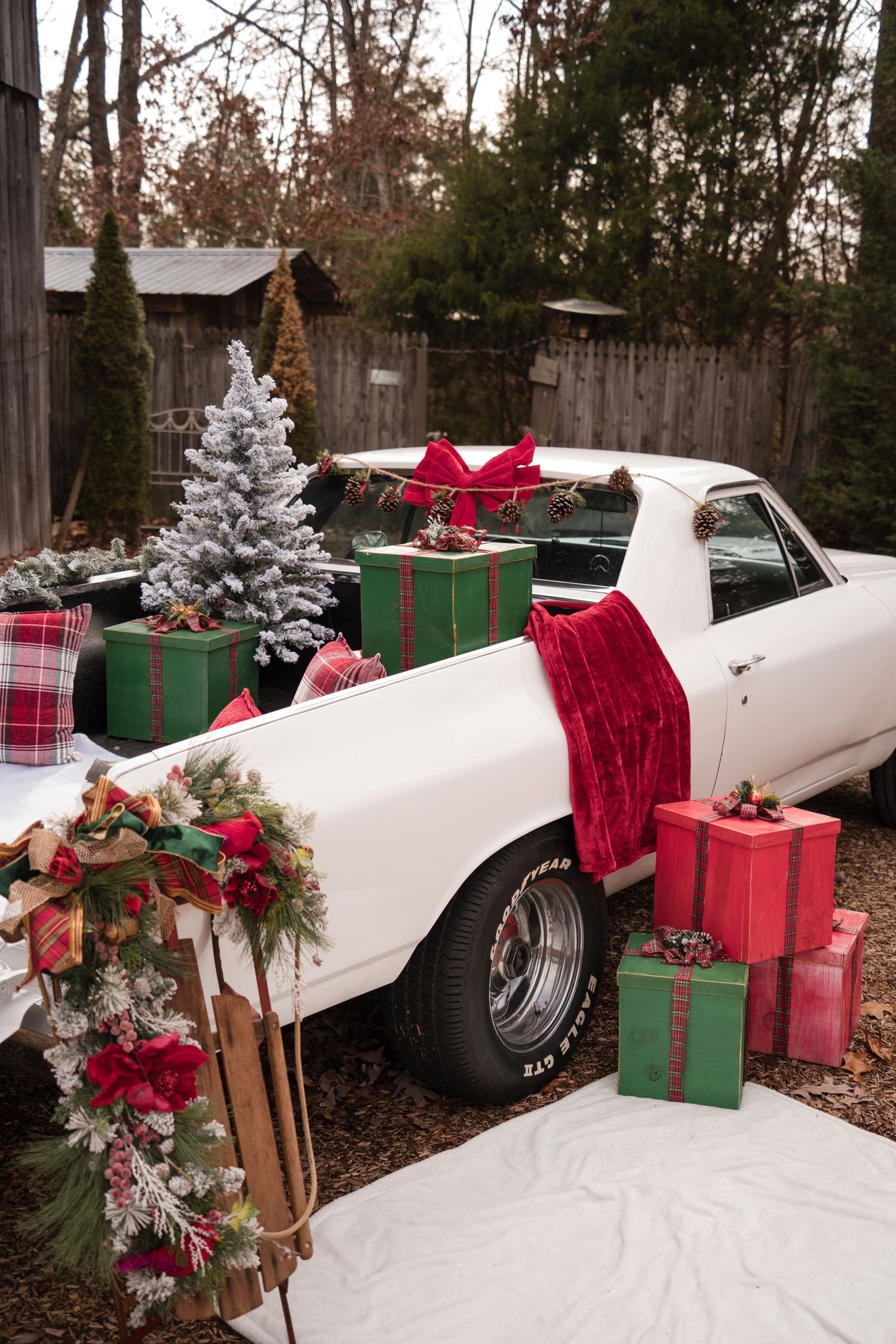 A vintage white car decorated for Christmas with red plaid bows, wrapped presents, and greenery in a winter outdoor setting.