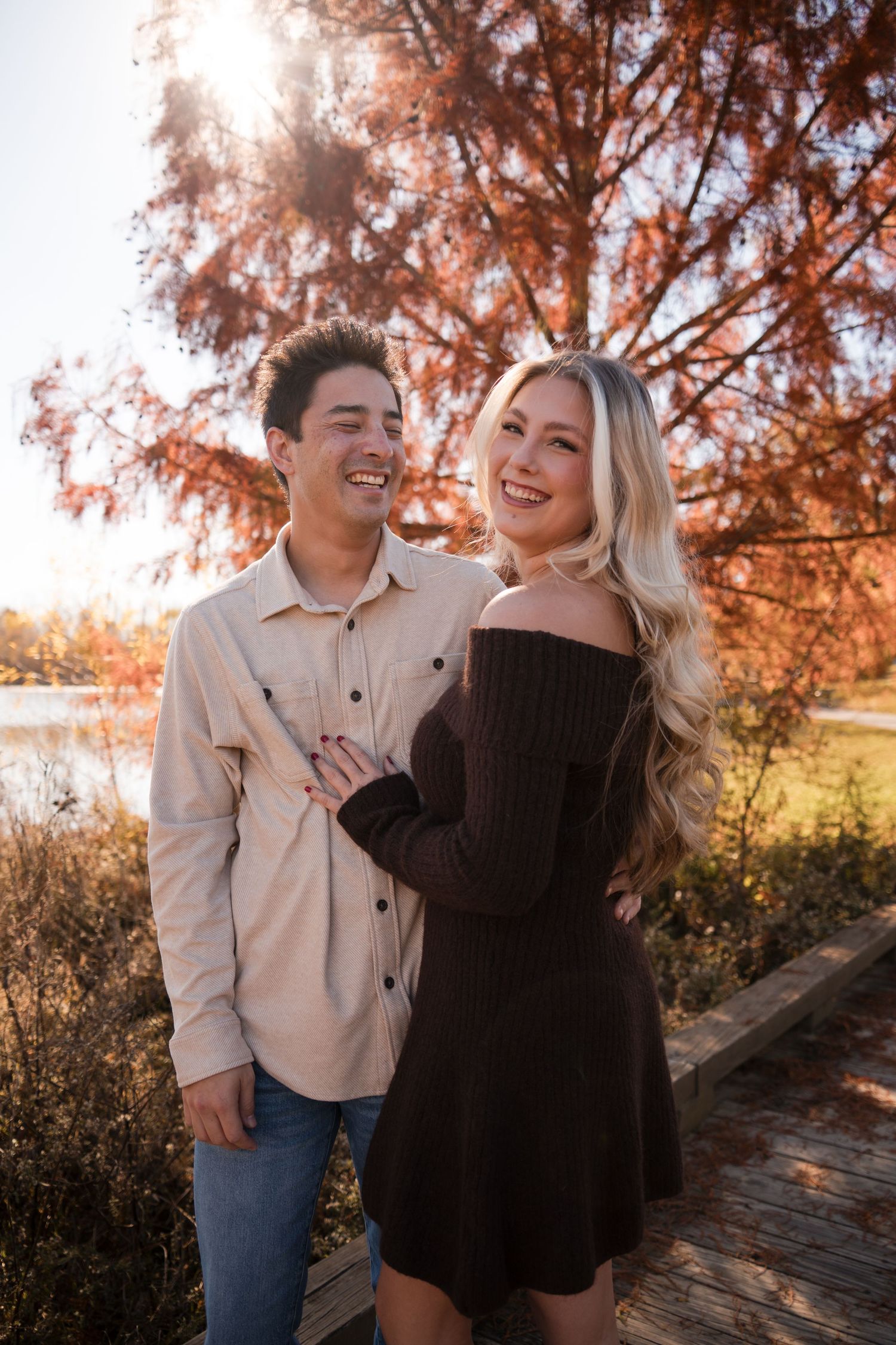 A couple laughs together during a sunset photoshoot on a wooden boardwalk surrounded by autumn foliage.