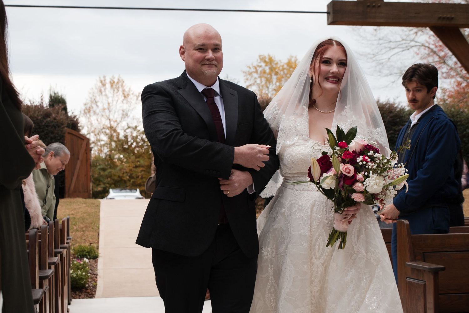 A bride in white walks down an aisle at an outdoor wedding ceremony during autumn.