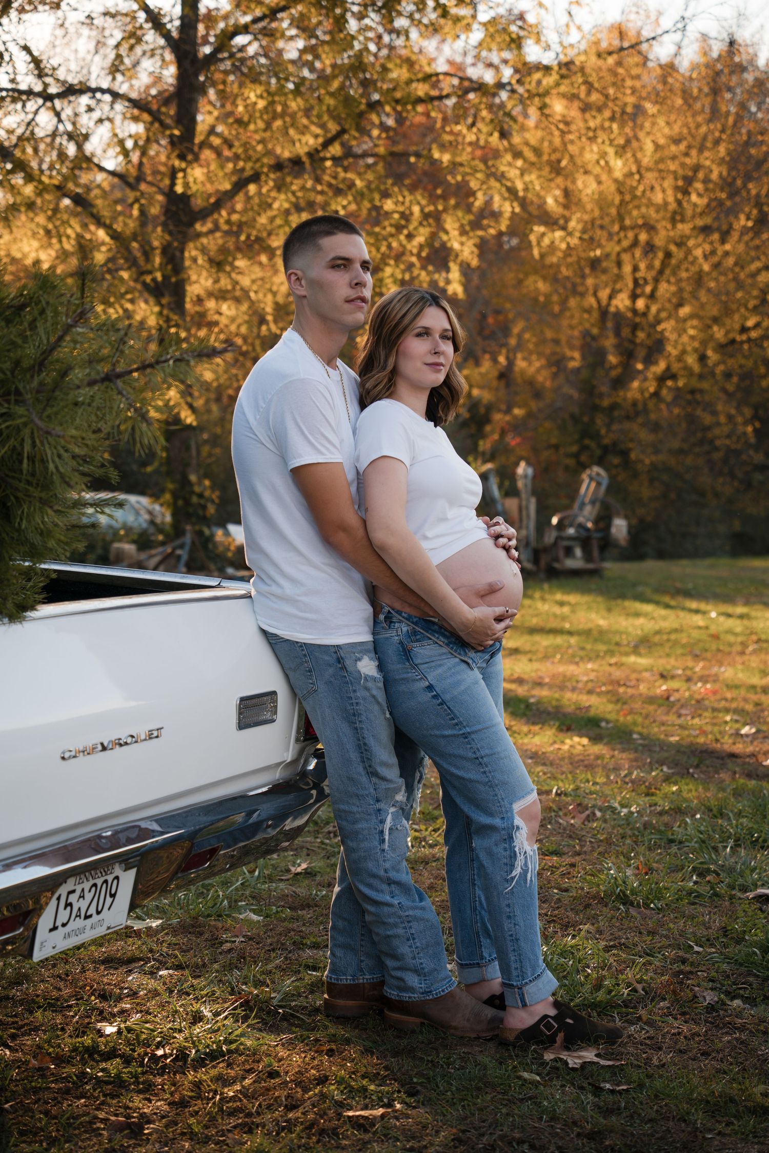 A couple in white t-shirts and jeans pose for a maternity photo by a vintage truck during golden hour in autumn.