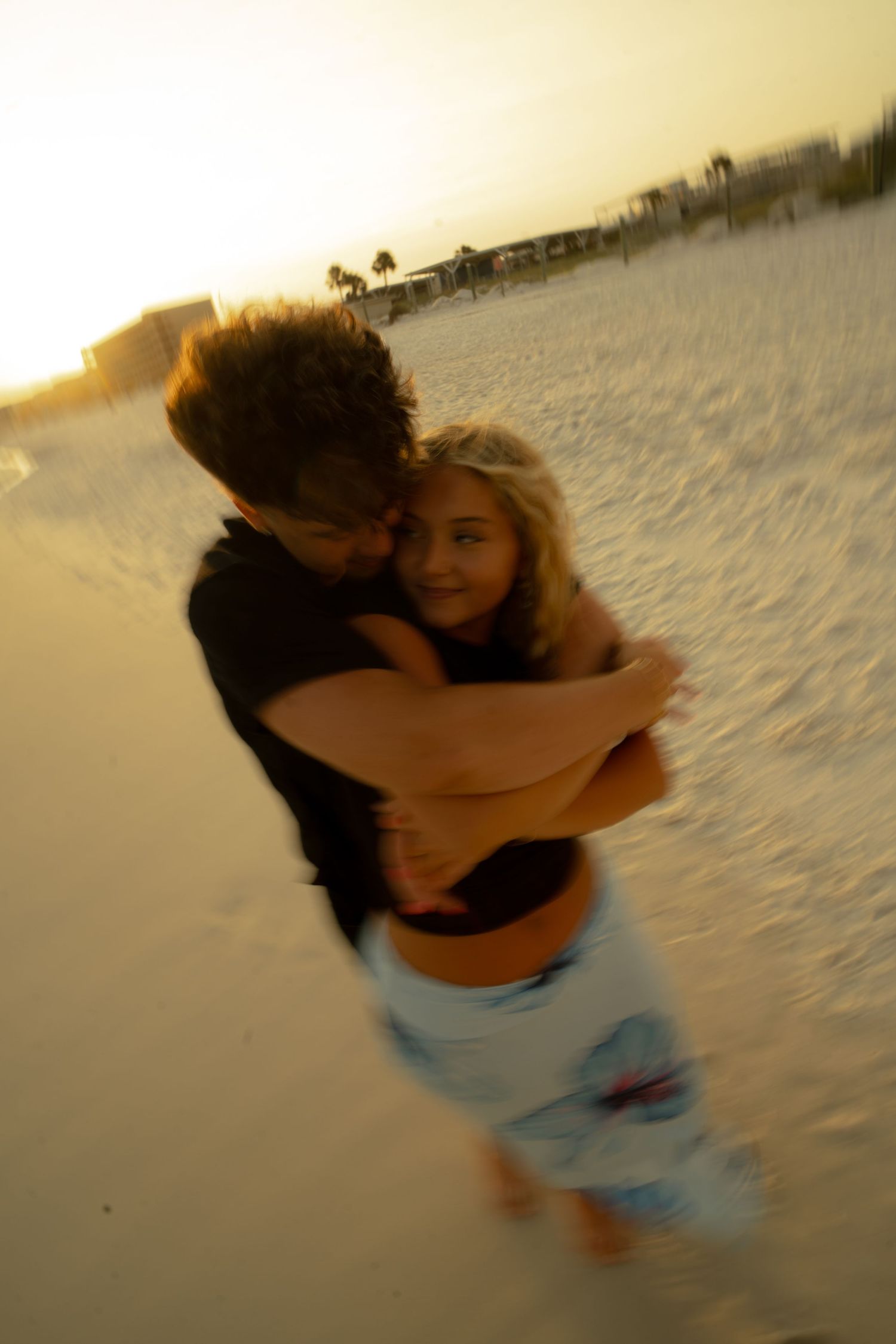 Couple embracing romantically on a sandy beach at golden sunset, man in black shirt hugging blonde woman in blue shorts.