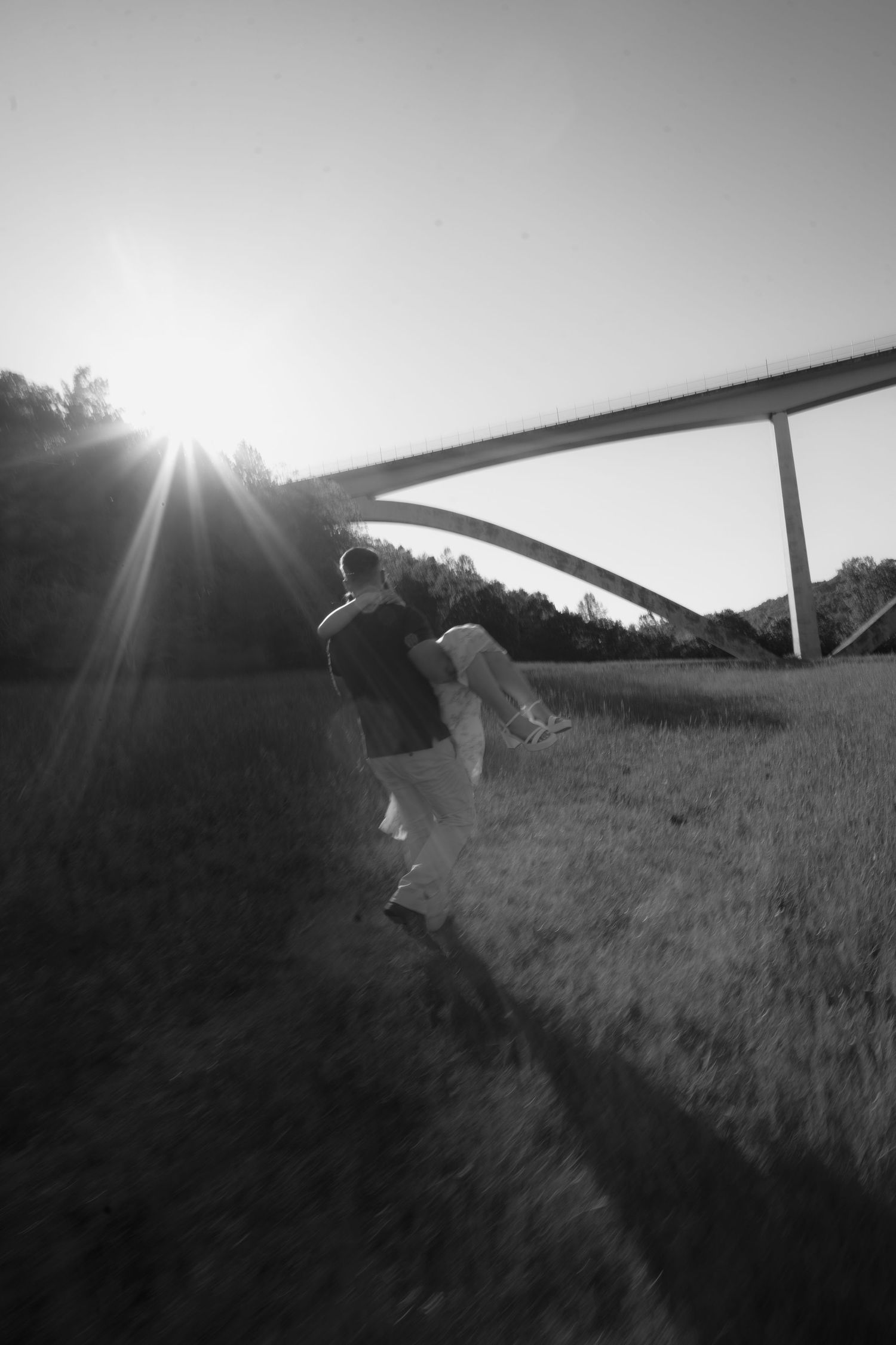 Black and white photo of a couple walking in a field at sunset with an arch bridge in the background.