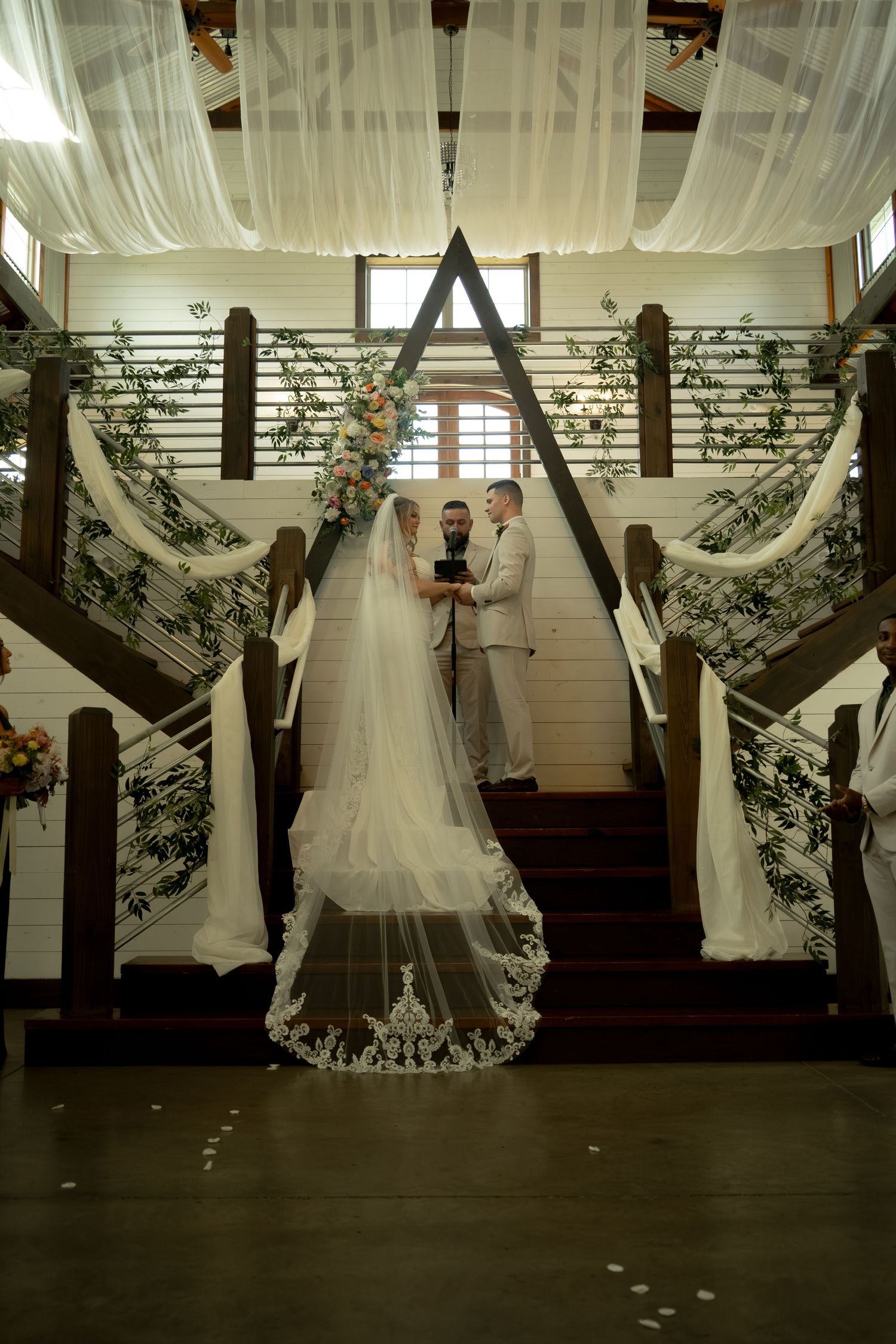 Bride and groom exchange vows at elegant staircase altar adorned with white draping and greenery during indoor wedding ceremony.