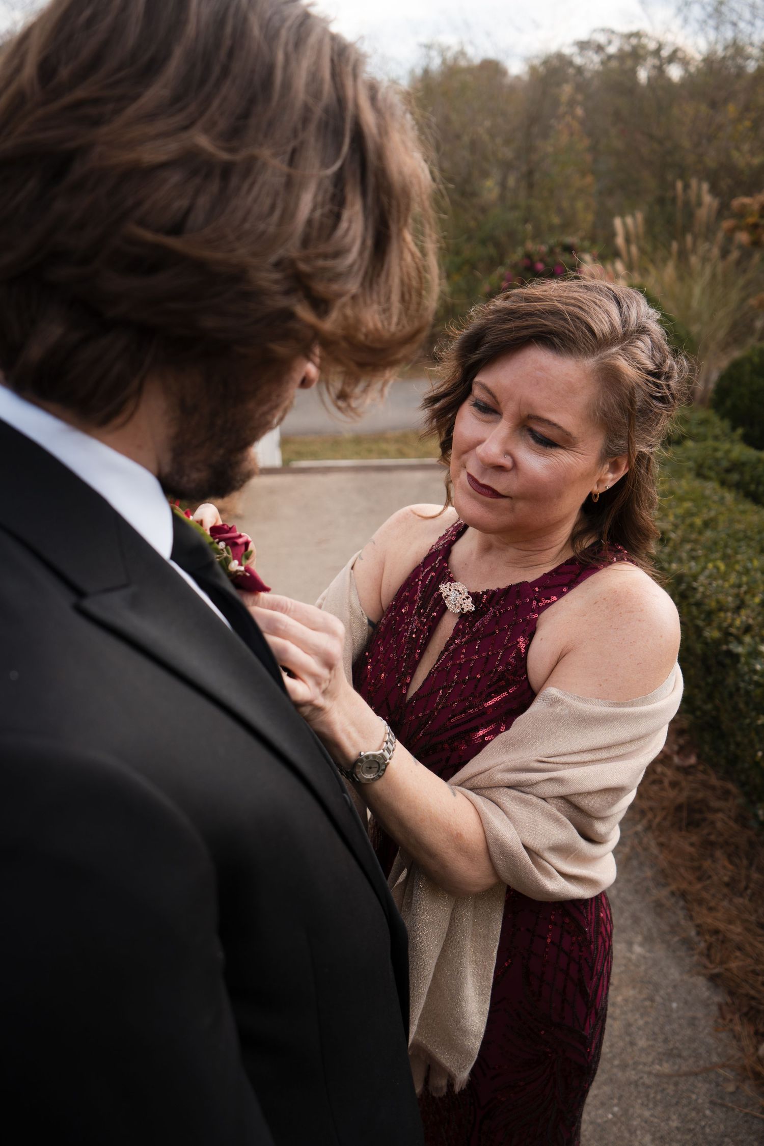 A couple shares an intimate moment outdoors while adjusting formal attire on a wooded path.