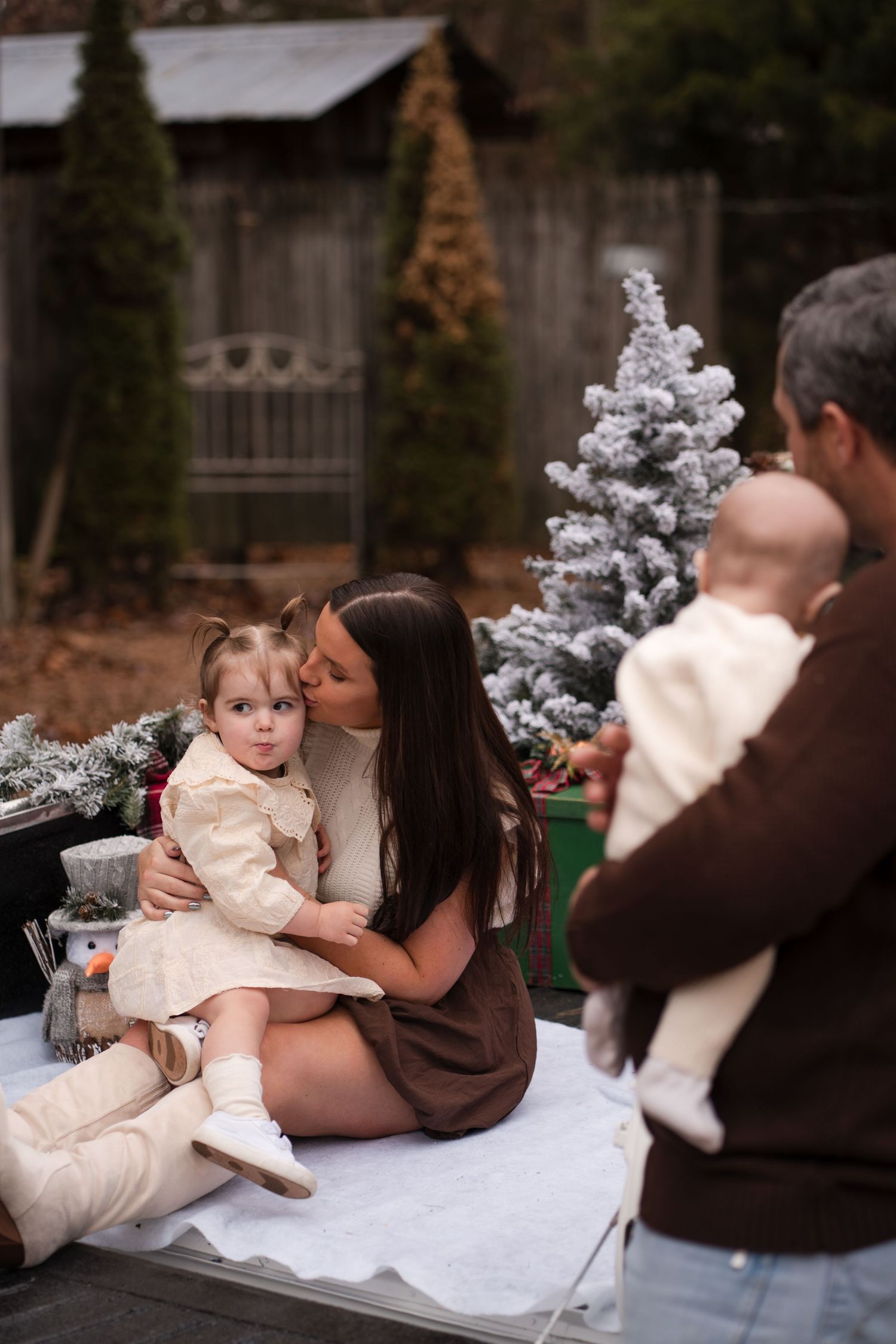 A family poses for holiday photos outdoors with a small decorated Christmas tree and white blanket.