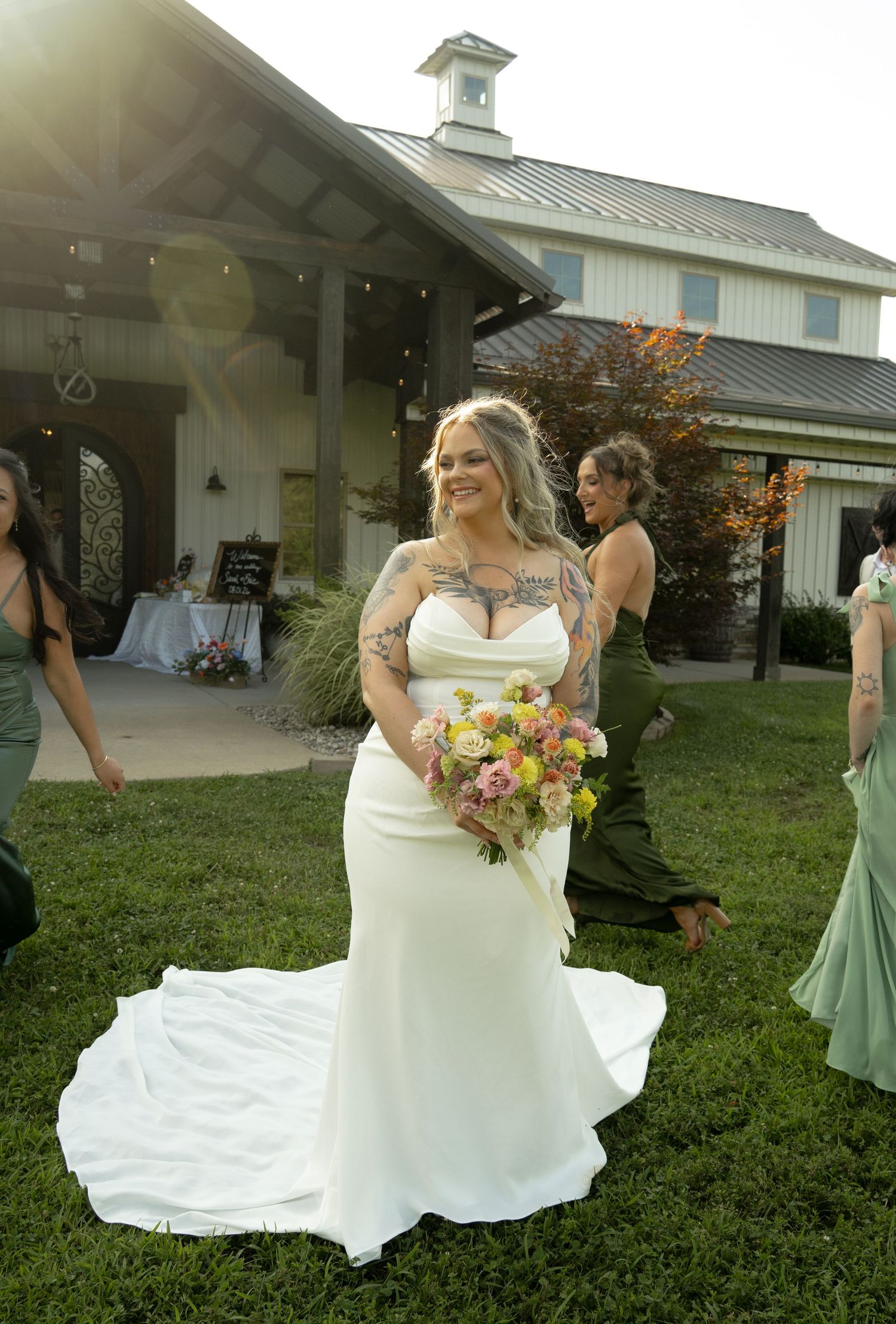 Tattooed bride in white mermaid gown holds colorful bouquet outdoors at rustic wedding venue with bridesmaids.