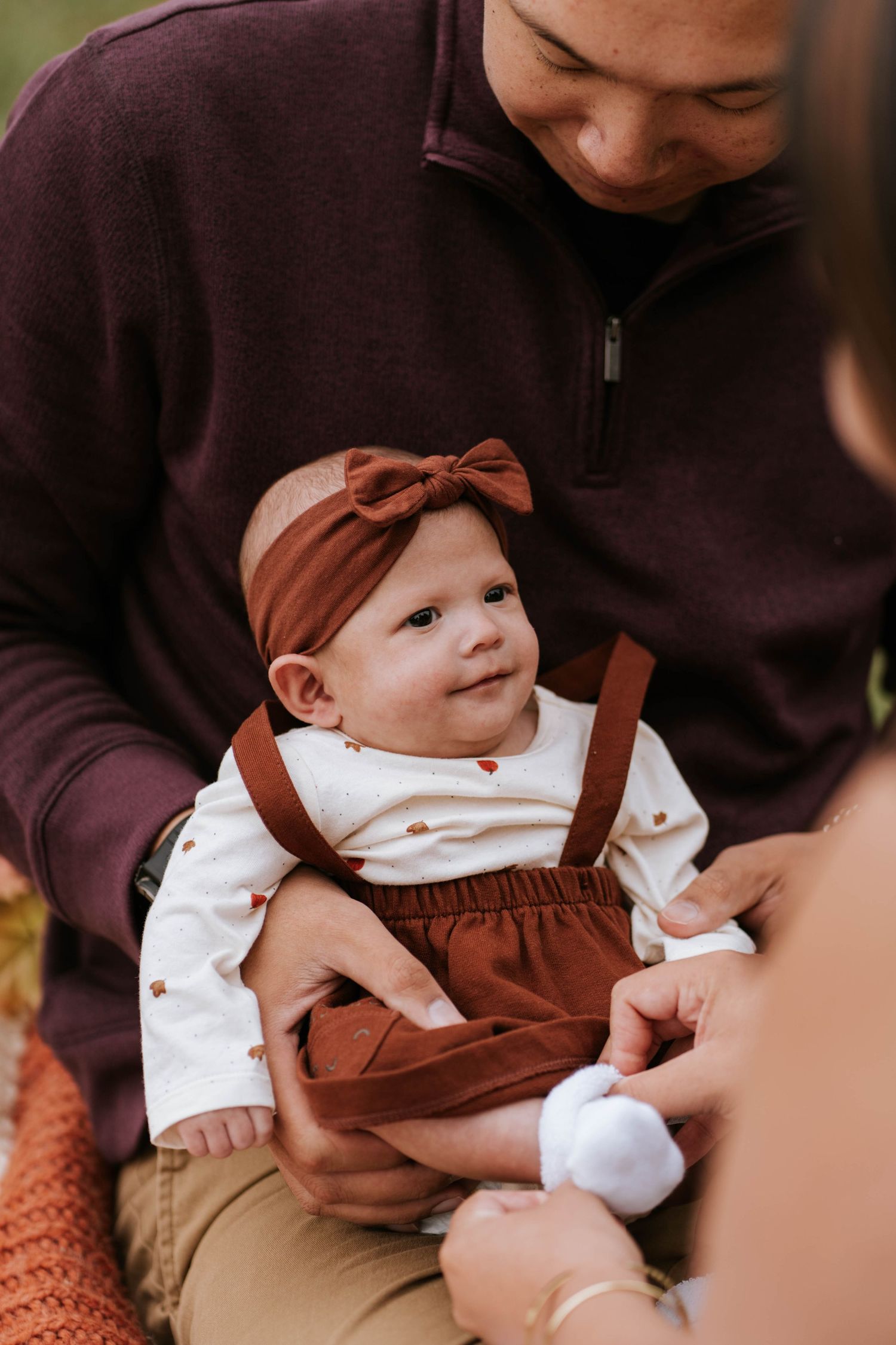 A baby wearing a brown bow headband and overalls sits contentedly while being held in a brown sweater.