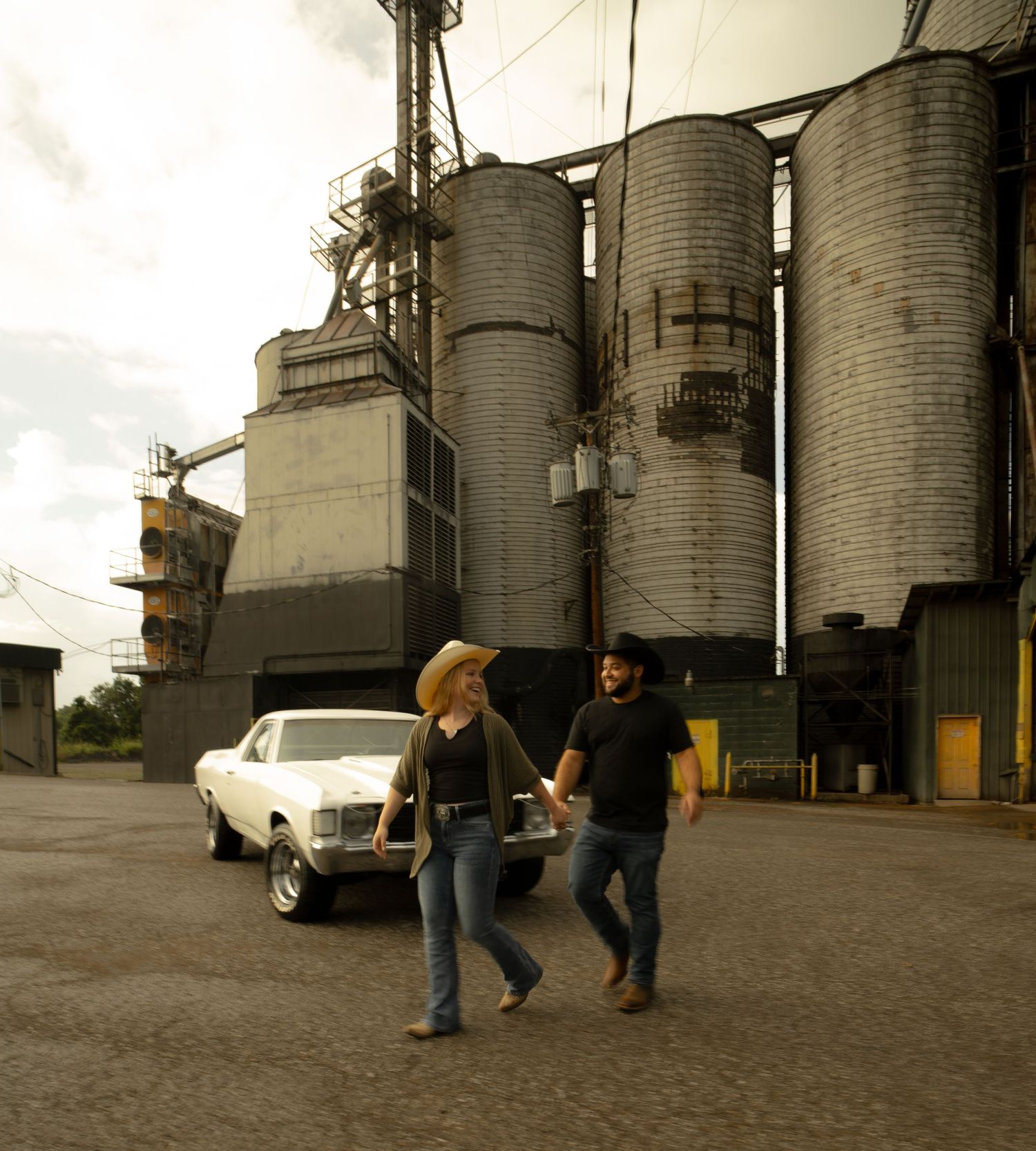 Couple holding hands walking past vintage white car and grain silos at industrial facility.