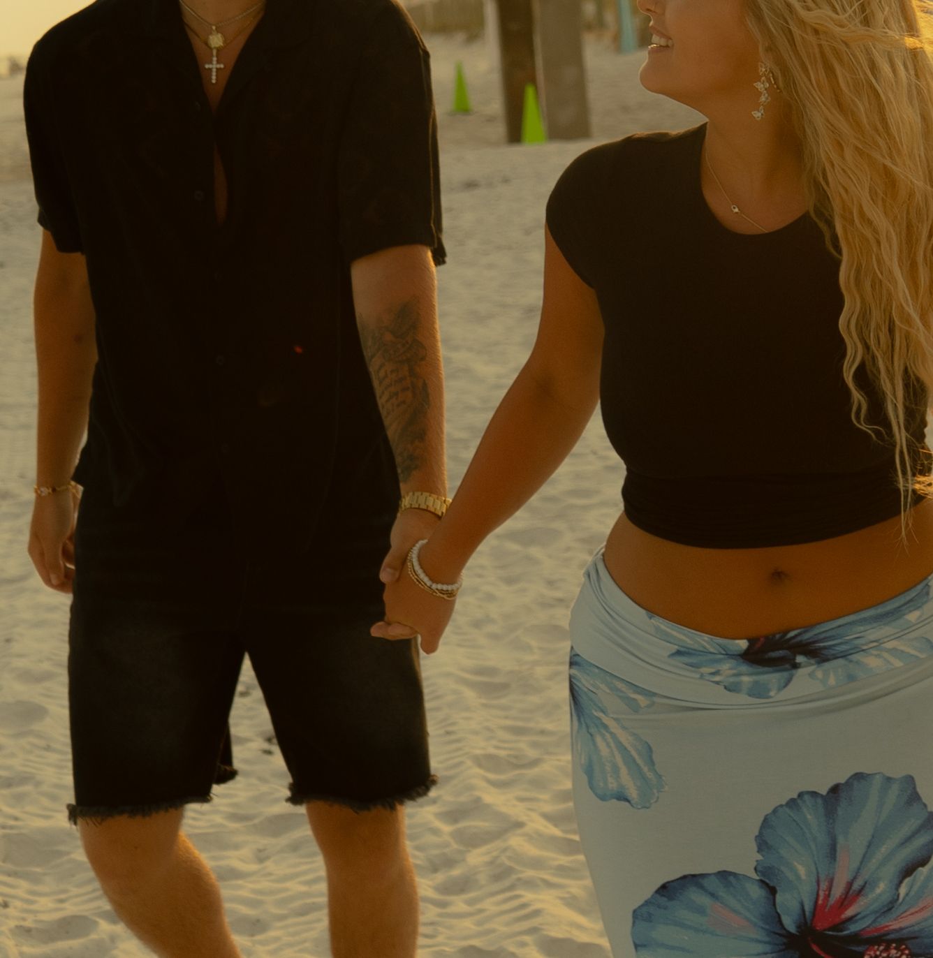 Couple holding hands on sunny beach, man with tattoos wearing black outfit, woman in floral skirt.