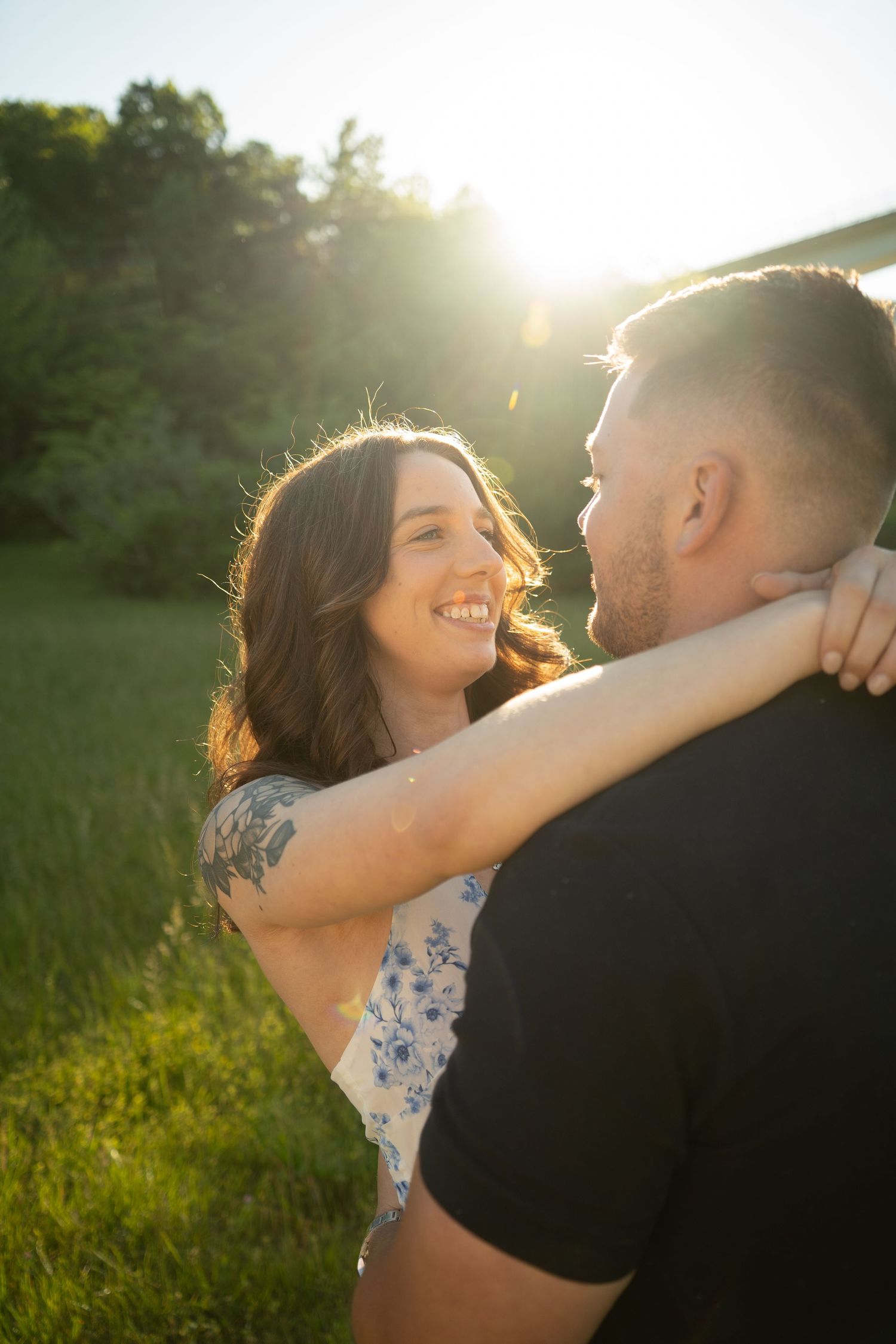 Couple embracing and smiling at each other in golden backlit sunlight in a lush green field.