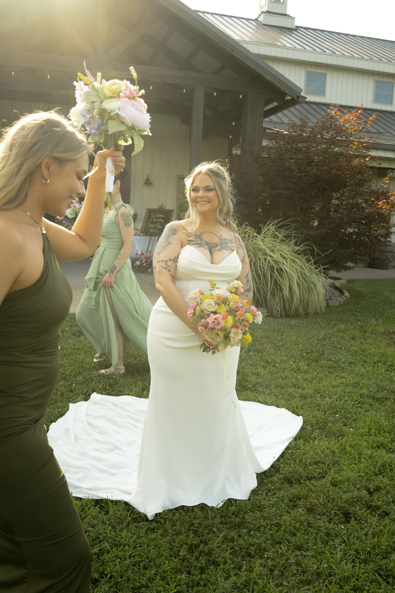 Bride in white gown holds colorful bouquet as bridesmaids in sage green dresses celebrate outdoors near a rustic venue.