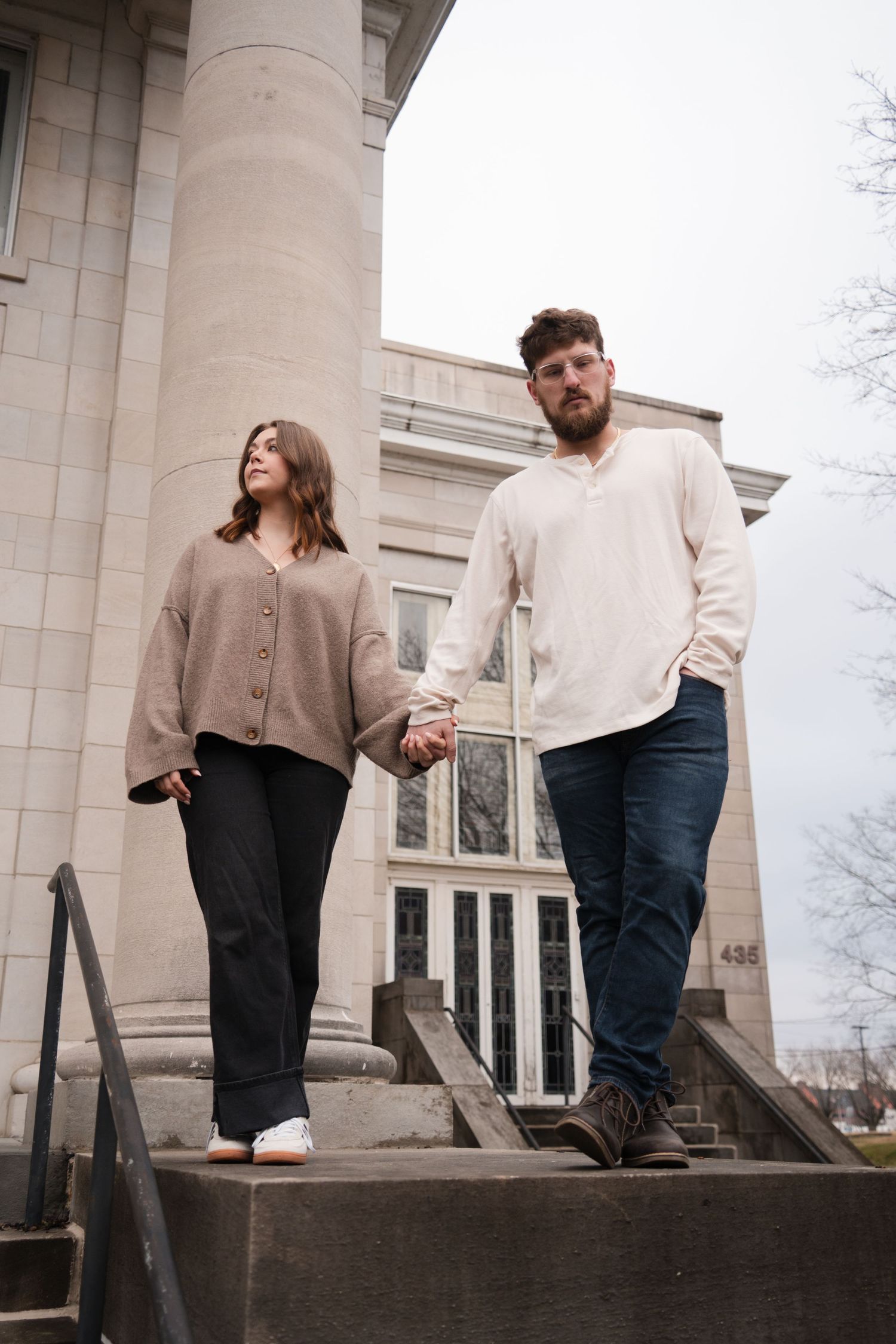 Couple holding hands walking down stone steps of a classic building, wearing casual neutral tones.