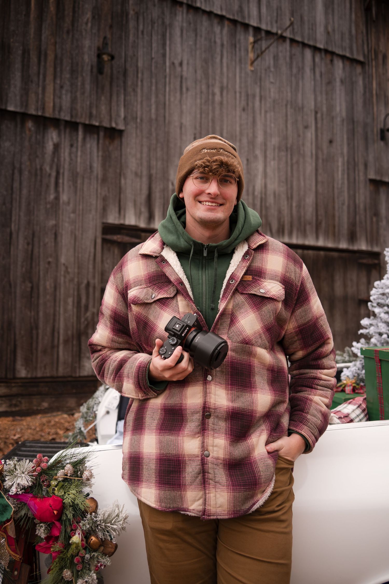A photographer in a brown knit cap, plaid flannel jacket and green hoodie holds a camera in front of a rustic barn.