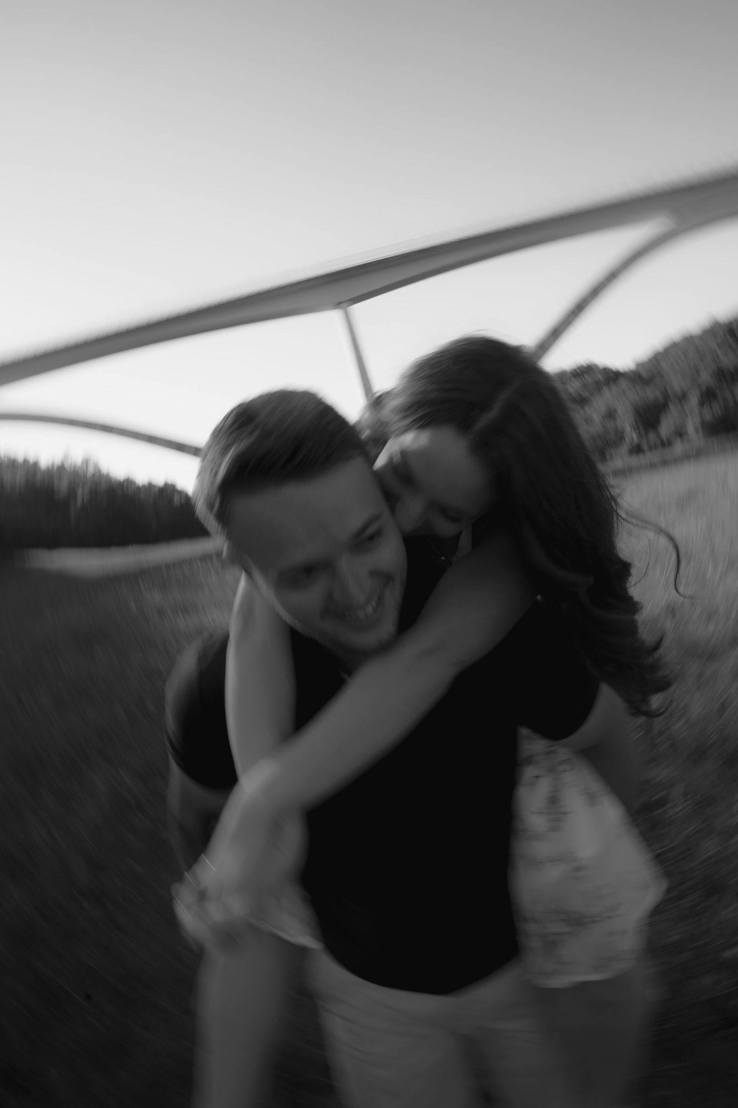 Black and white photo of a couple embracing and spinning outdoors near a bridge, captured with motion blur.