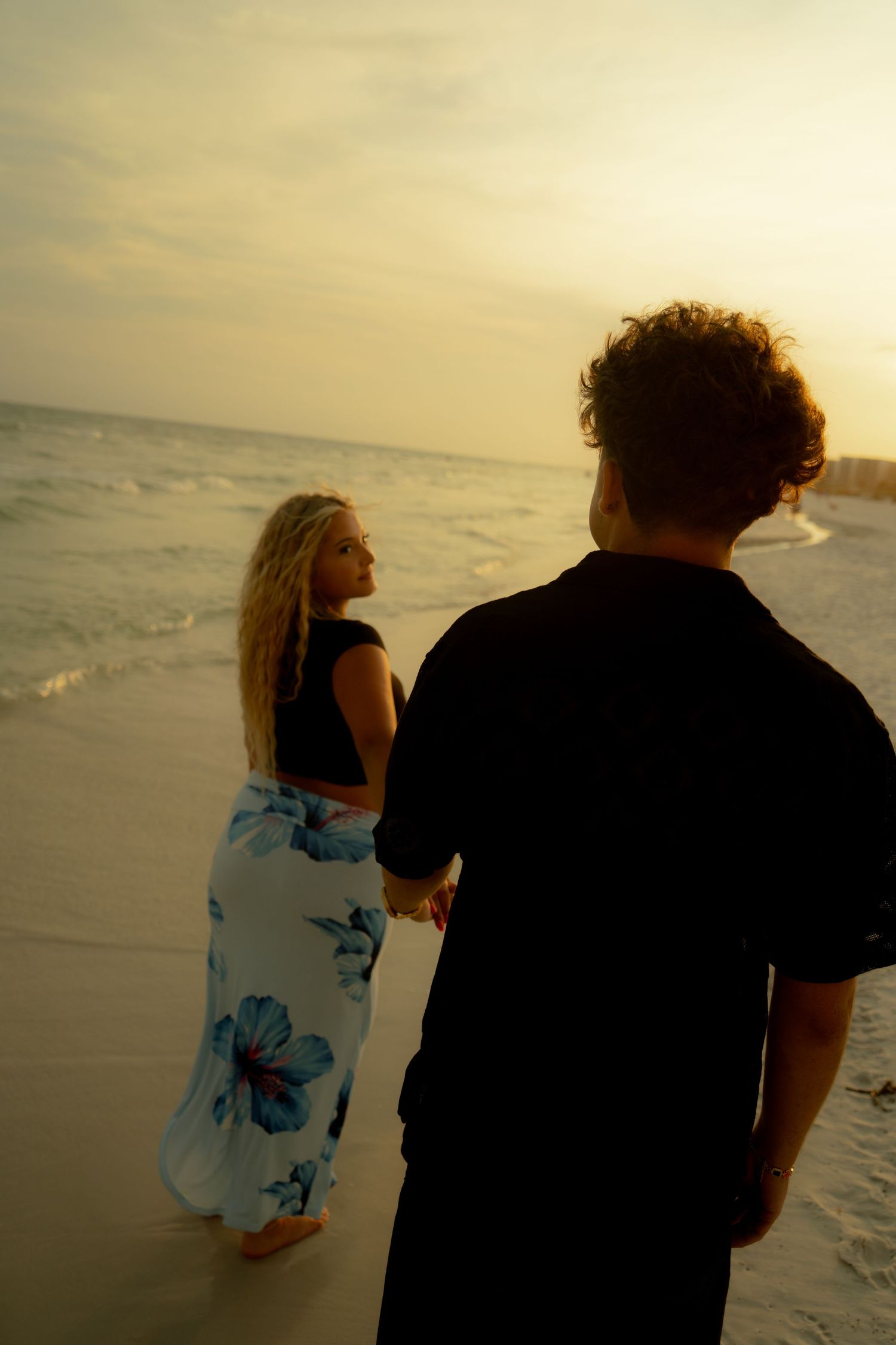 Couple holding hands on a sandy beach at sunset, woman in floral skirt looking back over her shoulder.