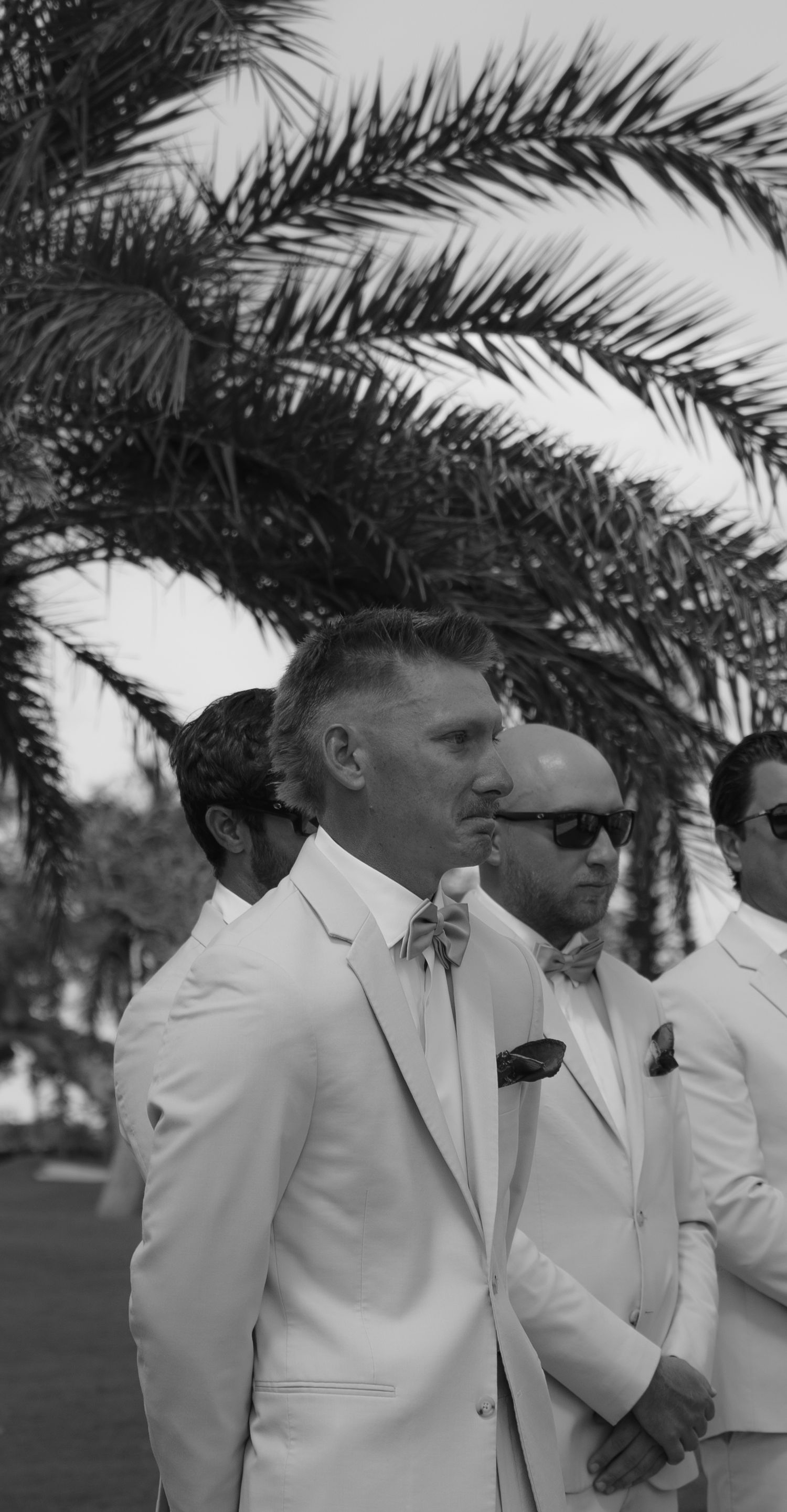 Black and white photo of groomsmen in white suits and bow ties standing outdoors near palm trees at a wedding.