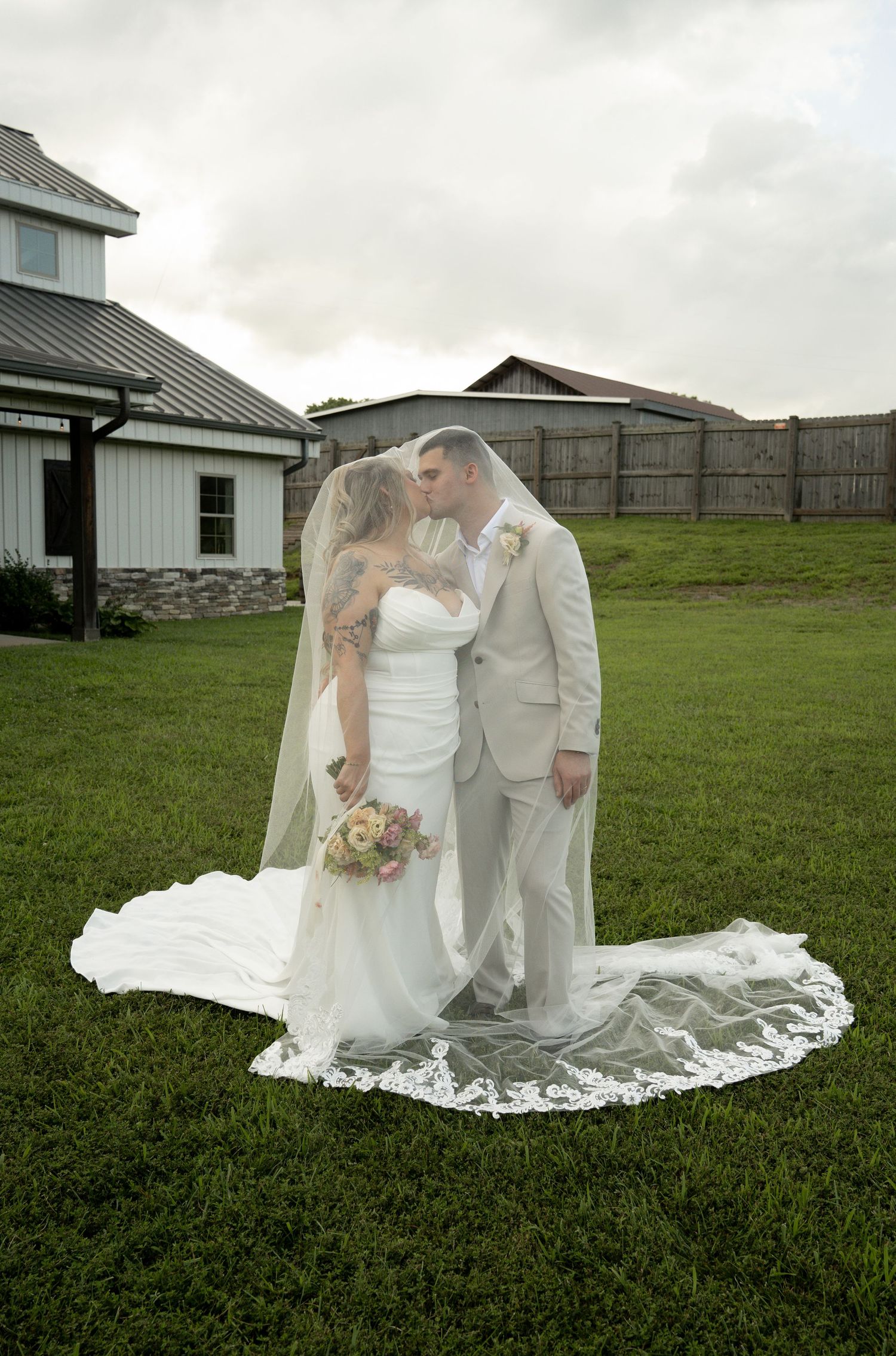 Bride and groom kissing under a flowing lace veil on green grass, with a white barn venue in the background.