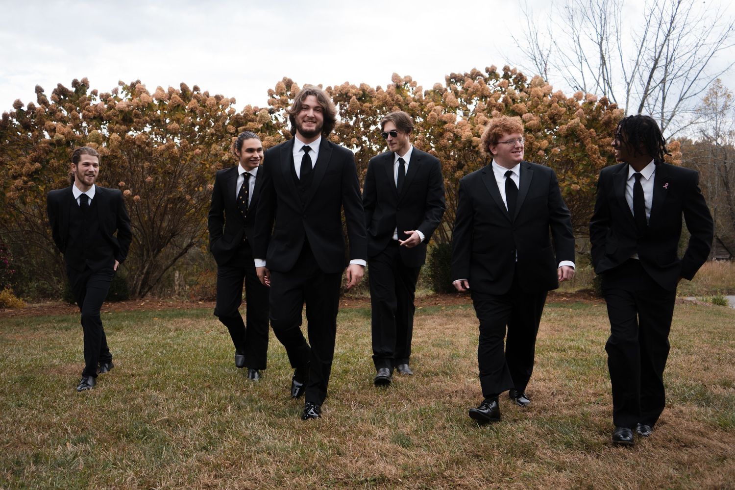 Groomsmen in black suits walk across a grassy field during an autumn wedding photo session.