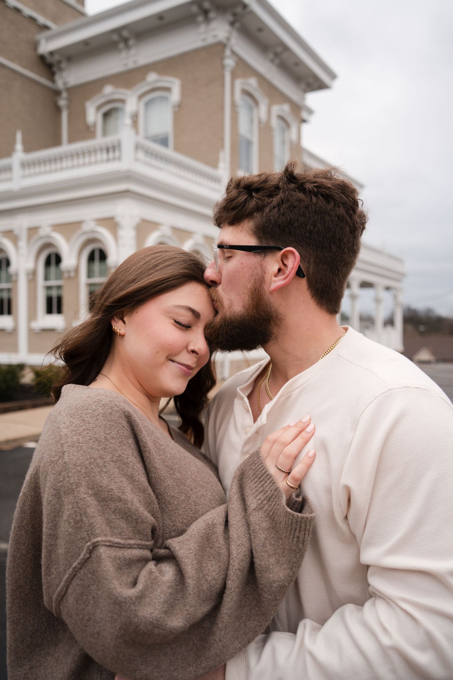 A couple shares a tender moment as a bearded man kisses a smiling woman on the forehead outside a historic building.