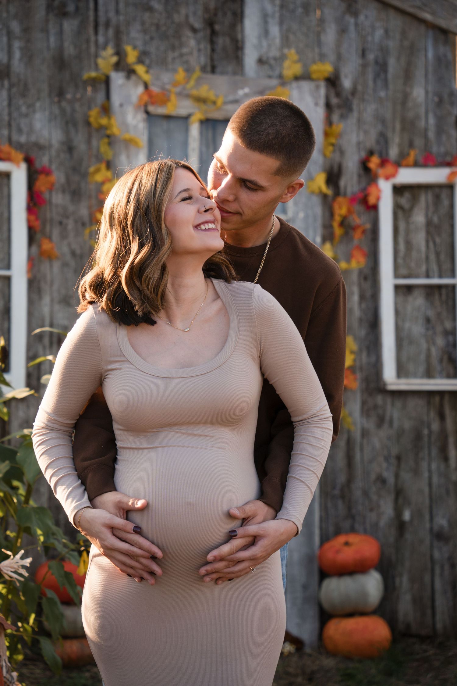 A couple shares tender moments in front of a rustic barn wall decorated with autumn pumpkins and foliage.